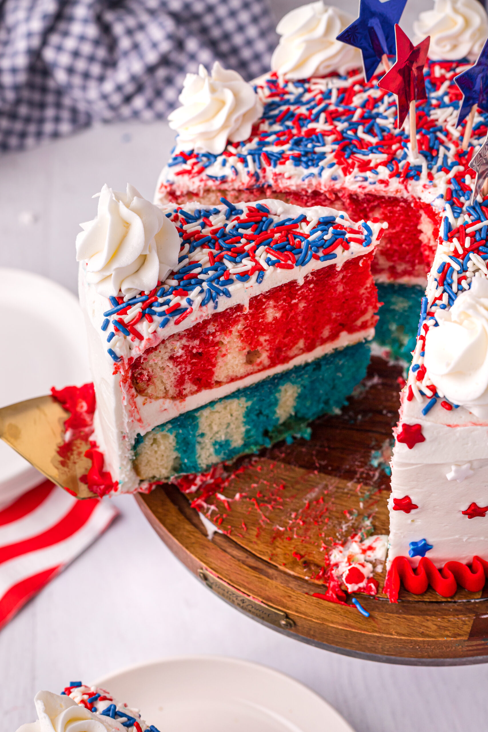Side view of 4th of July Cake on a wood cake stand with a slice being removed by a gold cake server.