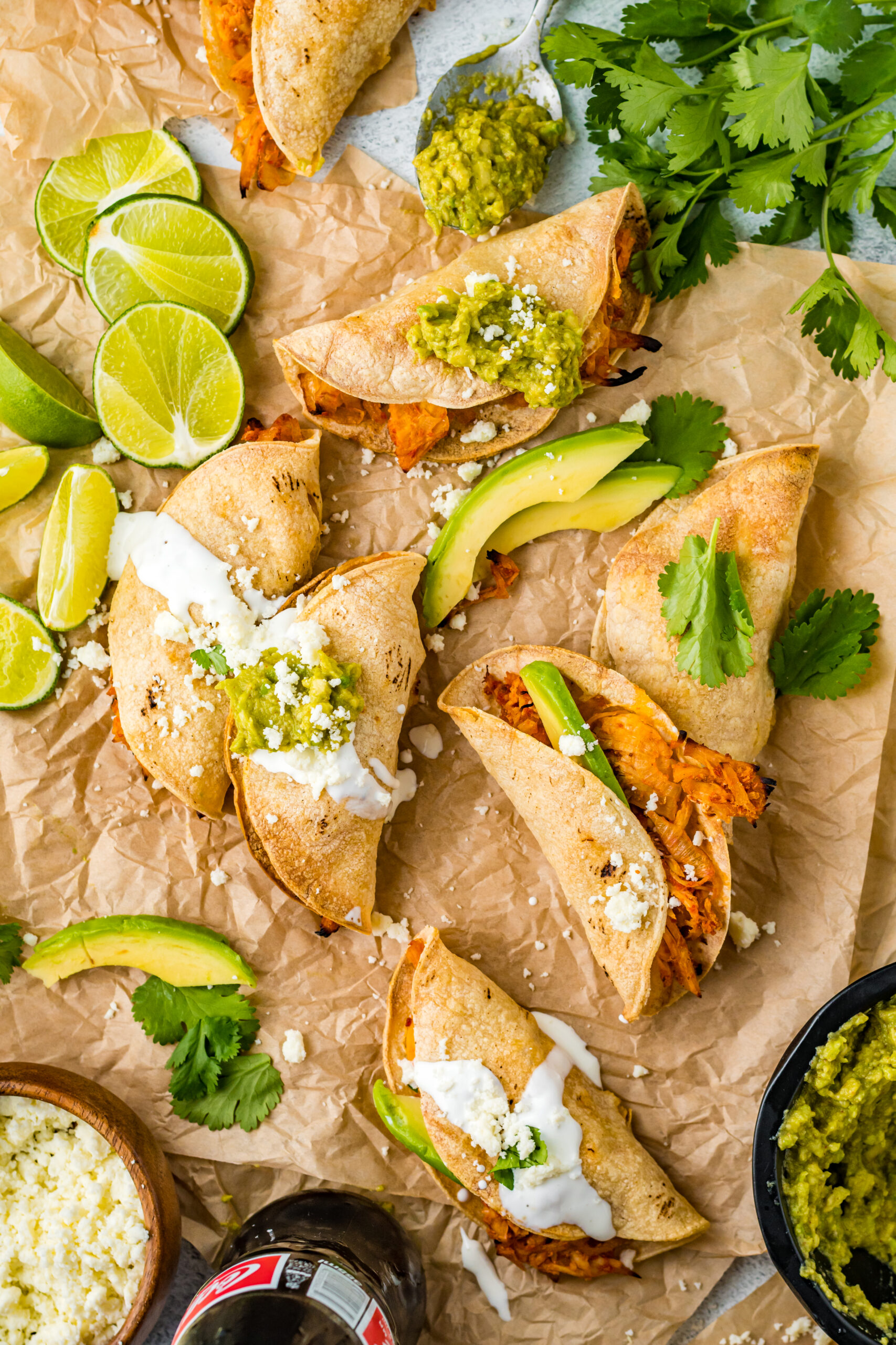 Overhead view of Crispy Tinga Tacos on a piece of parchment paper surrounded by lime and avocado slices.