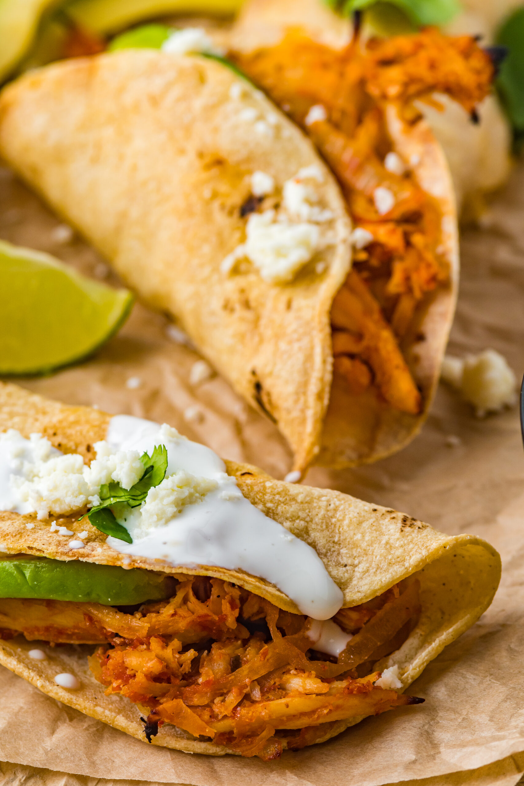 Close up view of Crispy Tinga Tacos on a piece of parchment paper surrounded by lime and avocado slices.