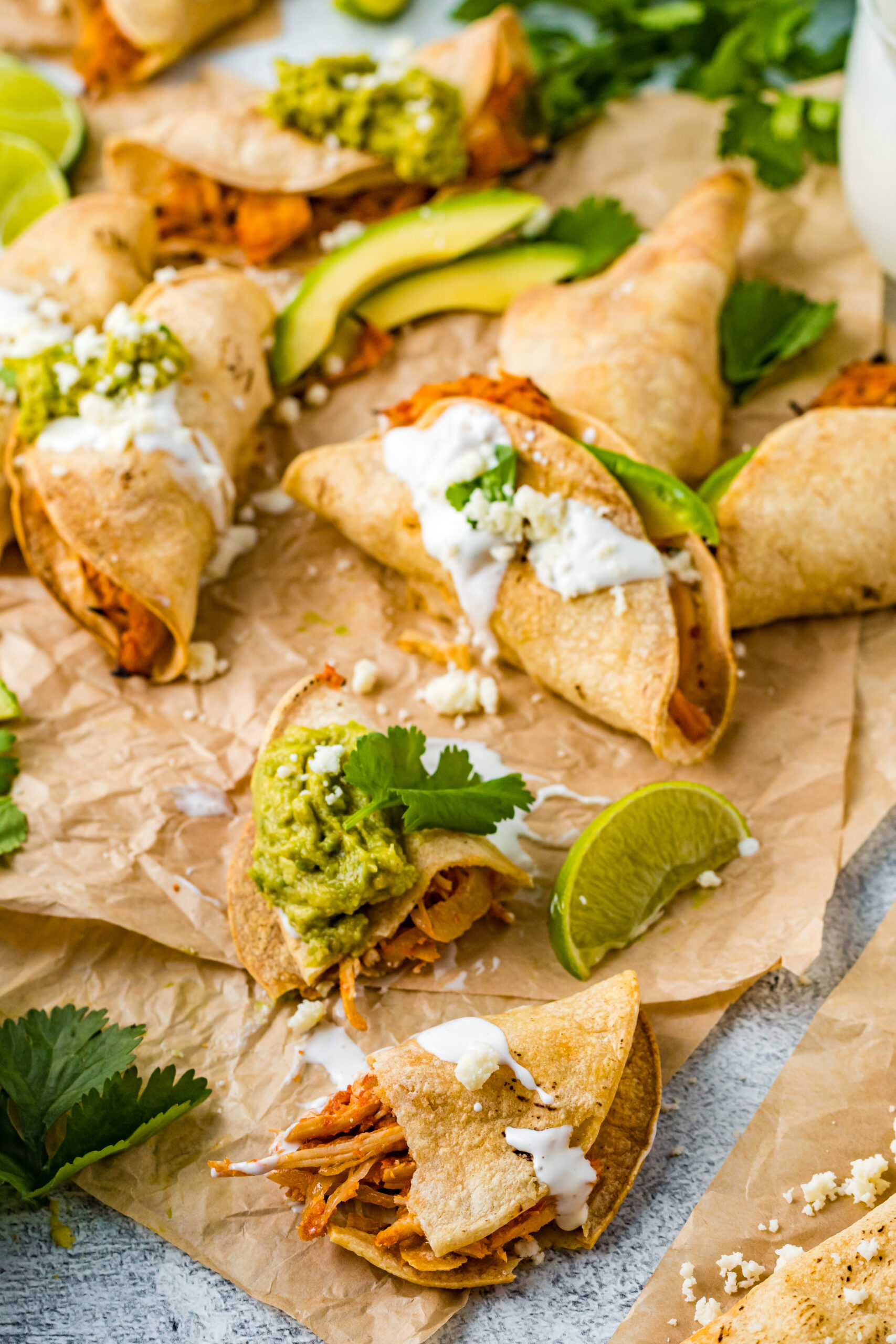 Front view of Crispy Tinga Tacos on a piece of parchment paper surrounded by lime and avocado slices with one taco split in half.
