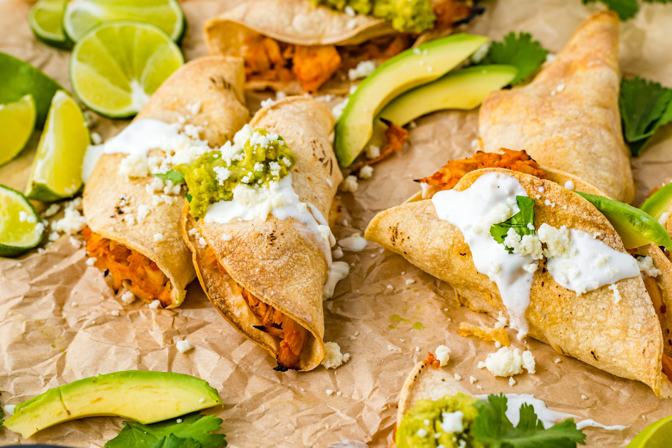 Front view of Crispy Tinga Tacos on a piece of parchment paper surrounded by lime and avocado slices.