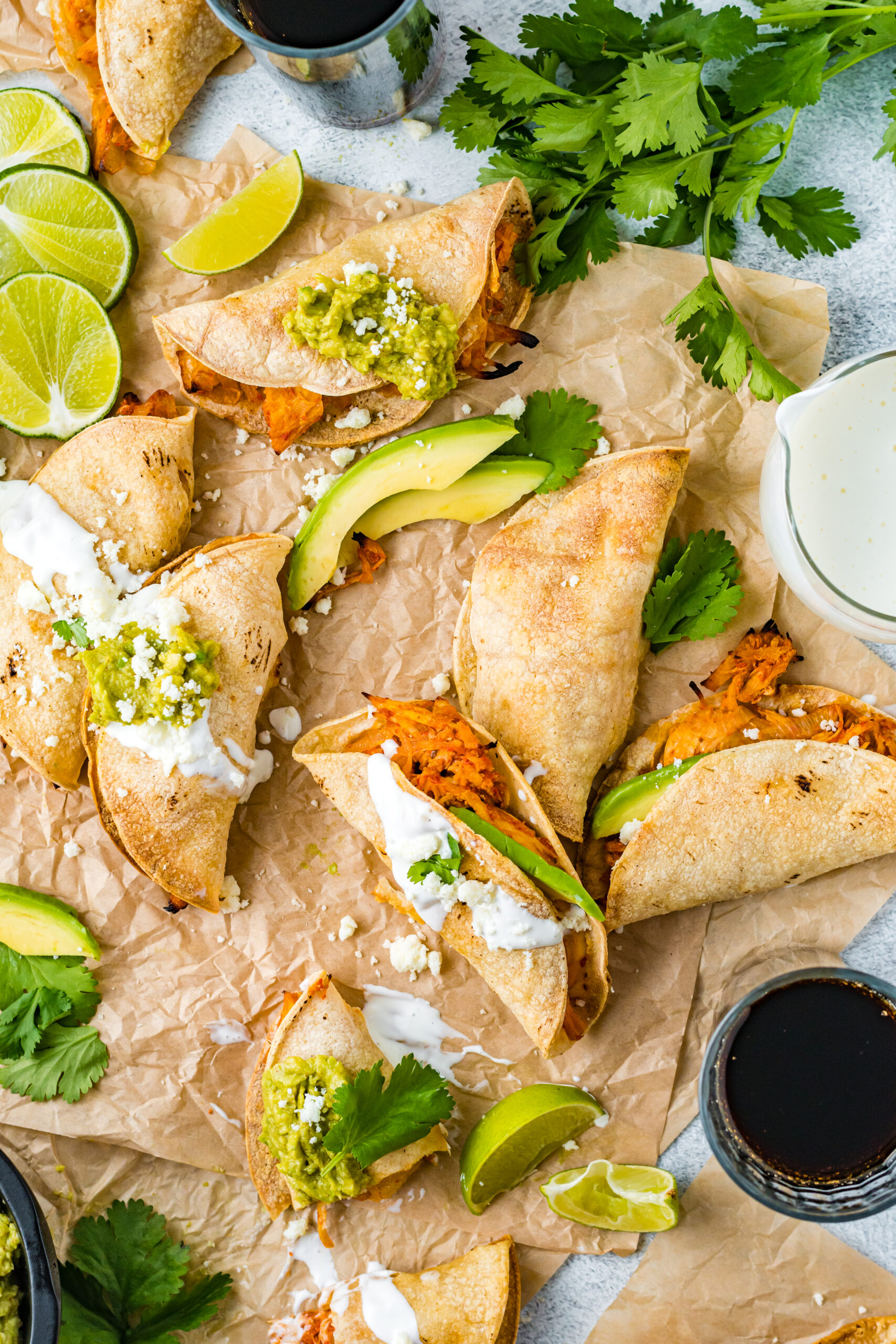 Overhead view of Crispy Tinga Tacos on a piece of parchment paper surrounded by lime and avocado slices.