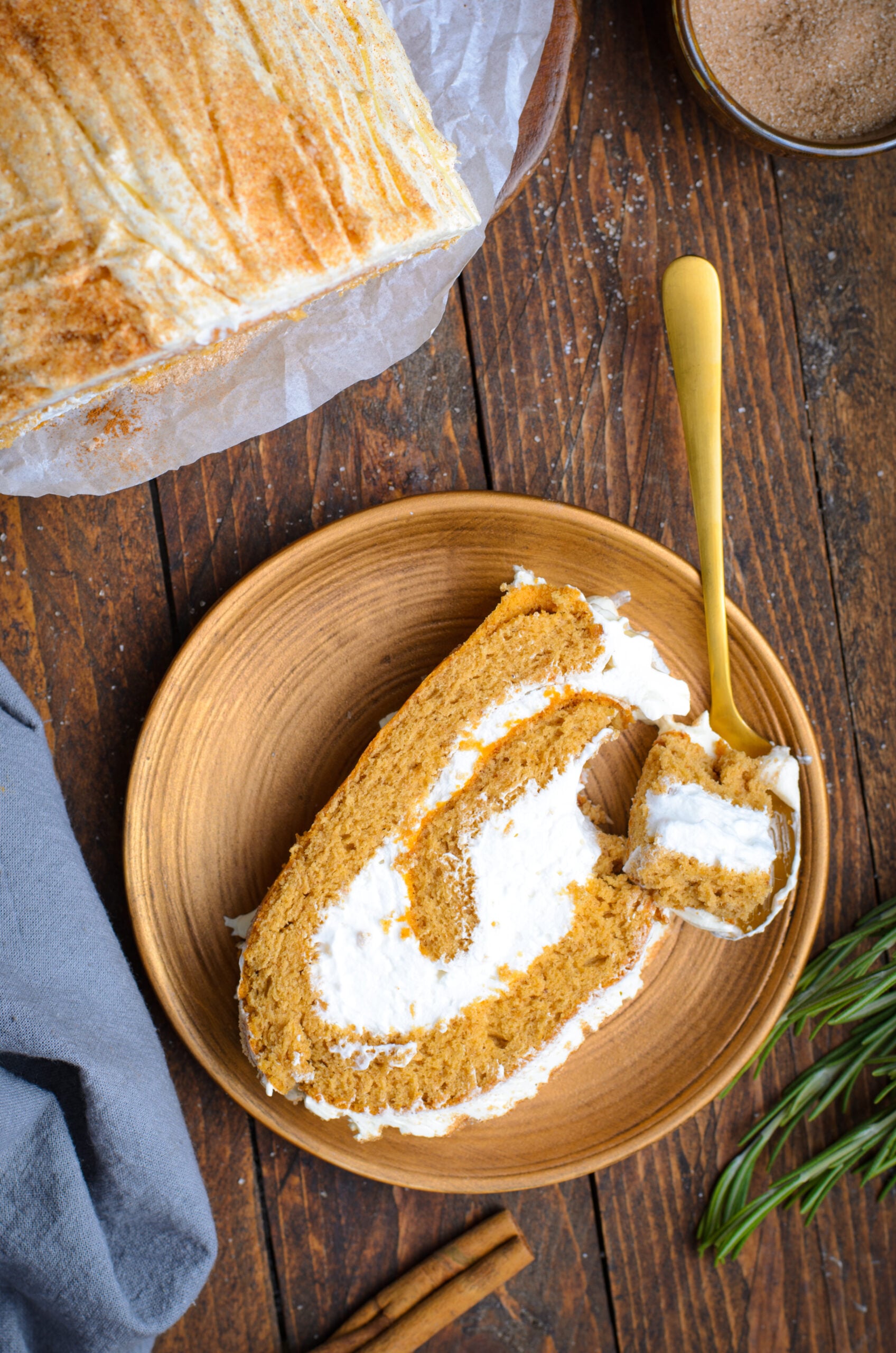 Overhead view of a slice of GIngerbread Cake Roll on a brown plate with a bite removed by a gold spoon.