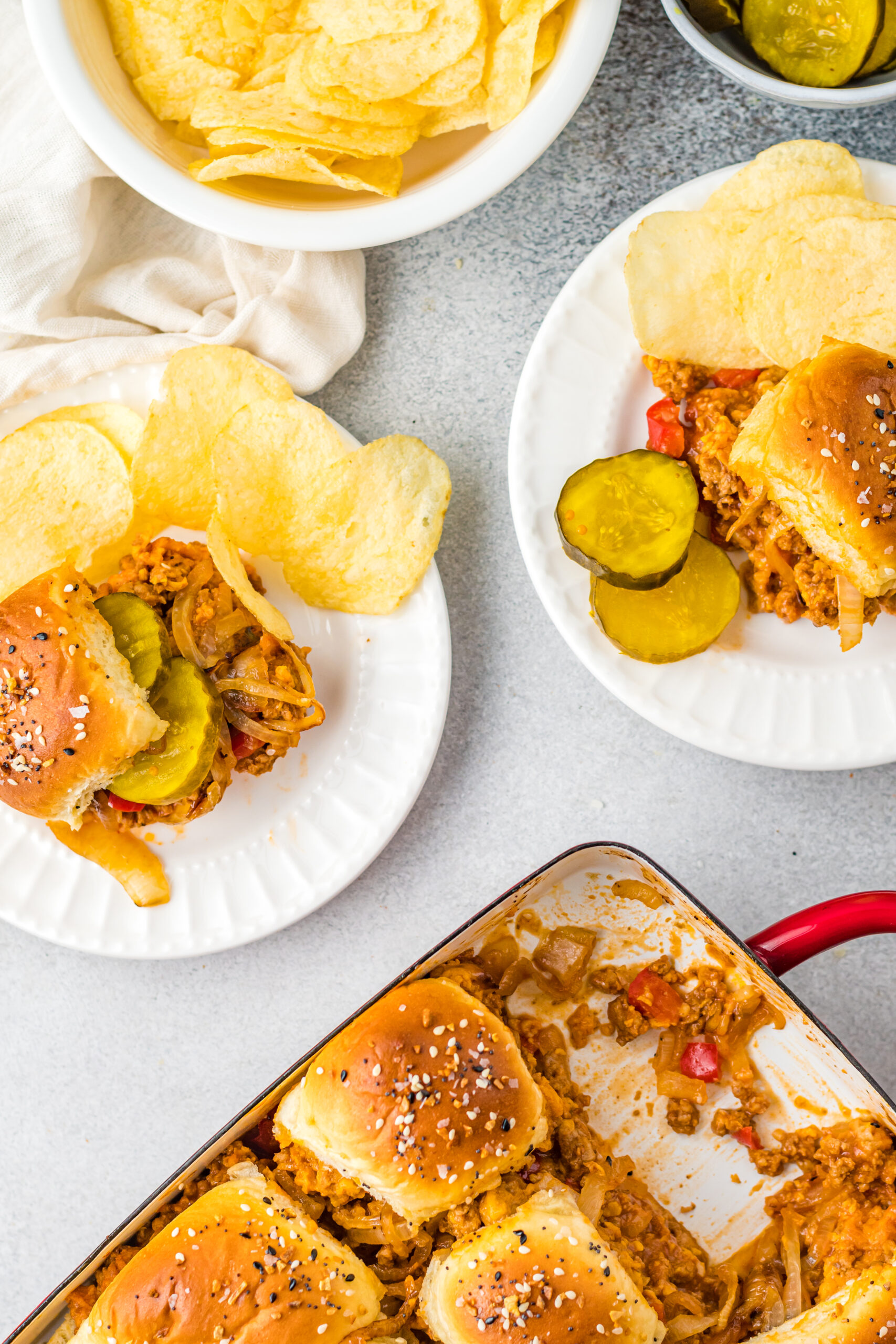 Overhead view of Cheesy Chicken Sloppy Joe Sliders on white plates served with pickle slices and chips.