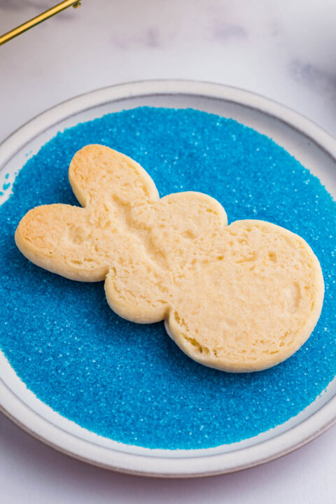 A bunny cookie being dipped in blue sprinkles.