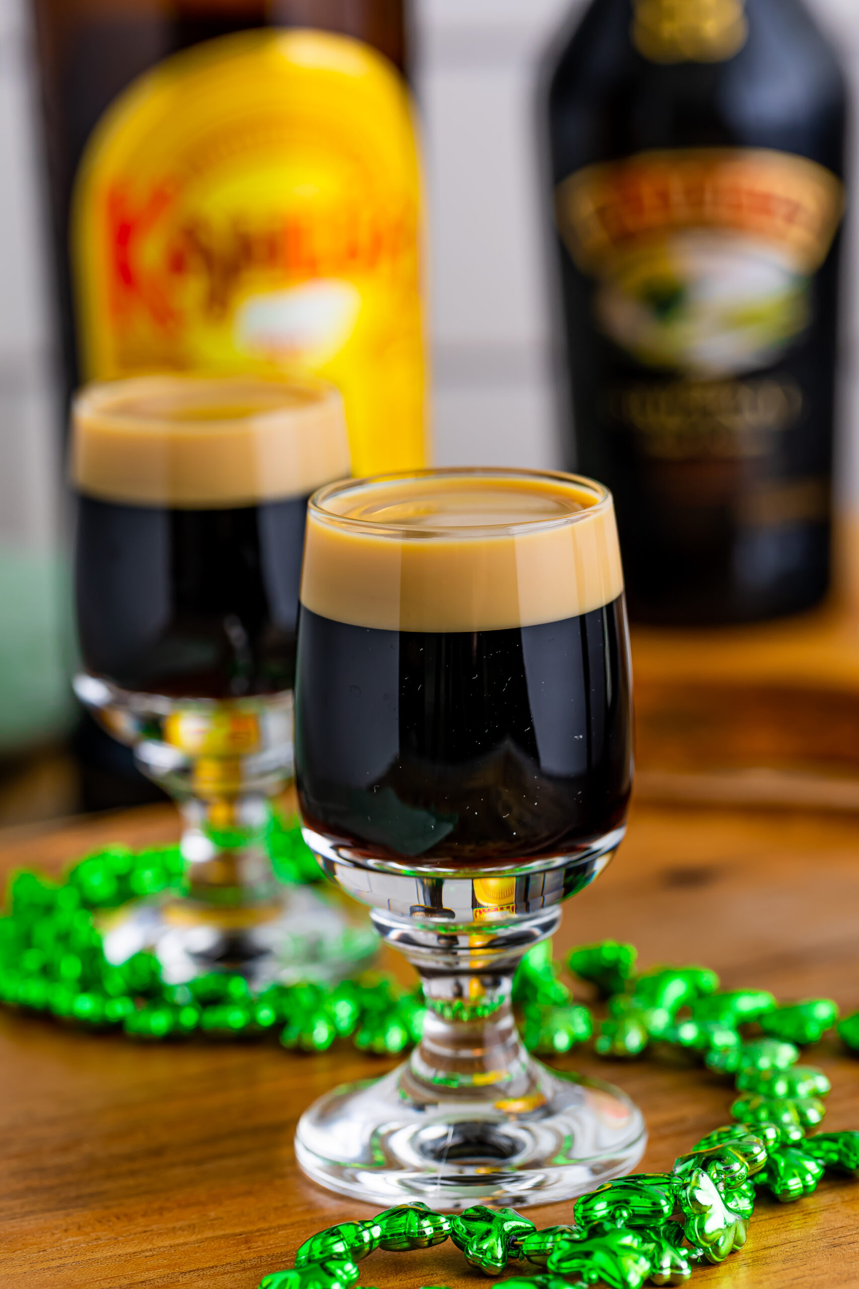Two Baby Guinness Shots on a wooden board with a green shamrock necklace. 