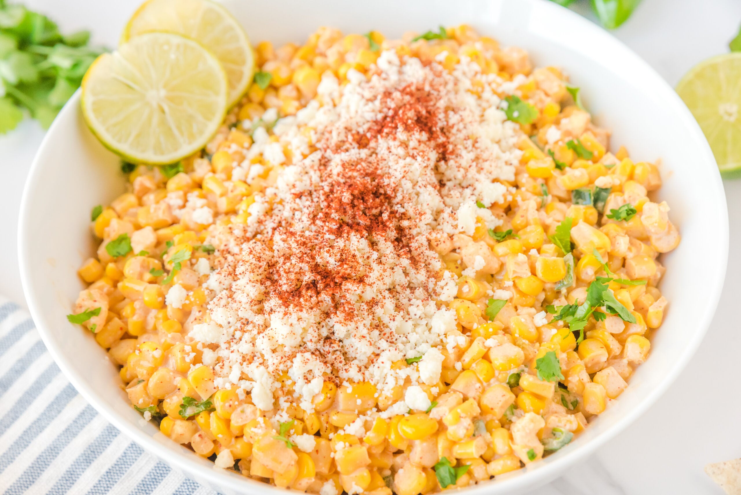 Mexican Street Corn Salad in a large white serving bowl, topped with feta cheese and sliced limes.