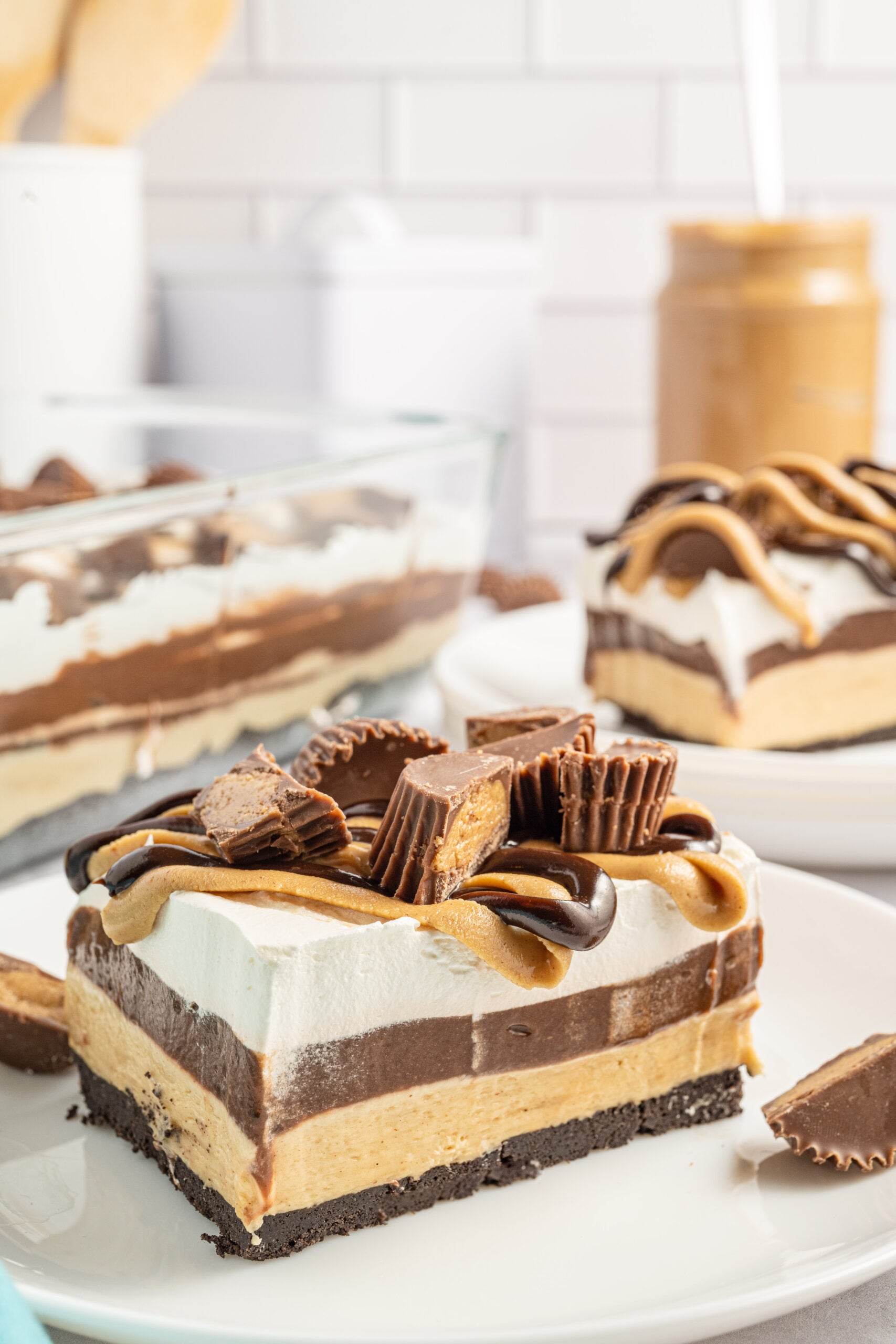 Two pieces of chocolate peanut butter lasagna on two white plates with a jar of peanut butter in the background. 
