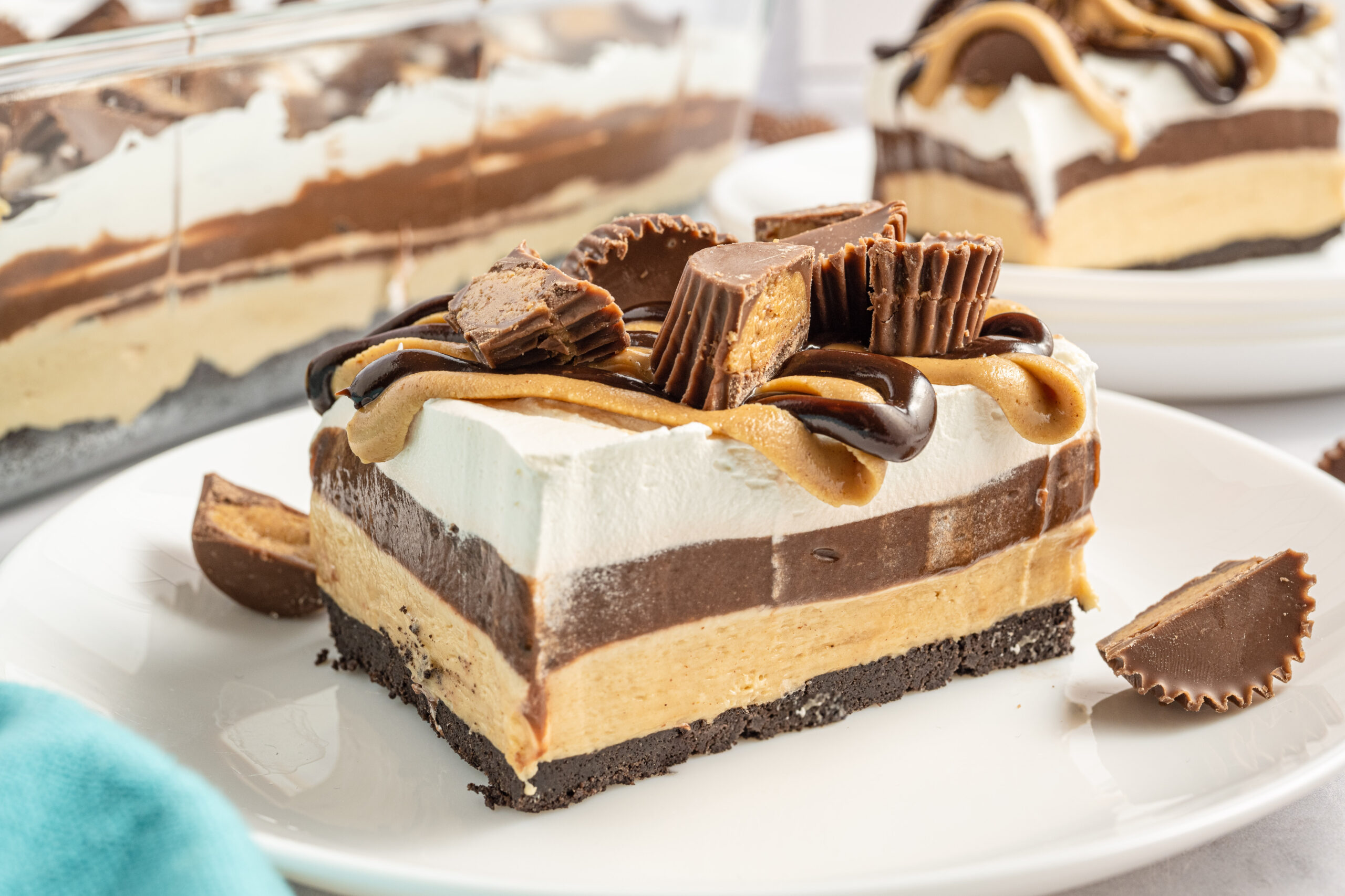 A piece of chocolate peanut butter lasagna on a white plate with Reese's cups in the background. 