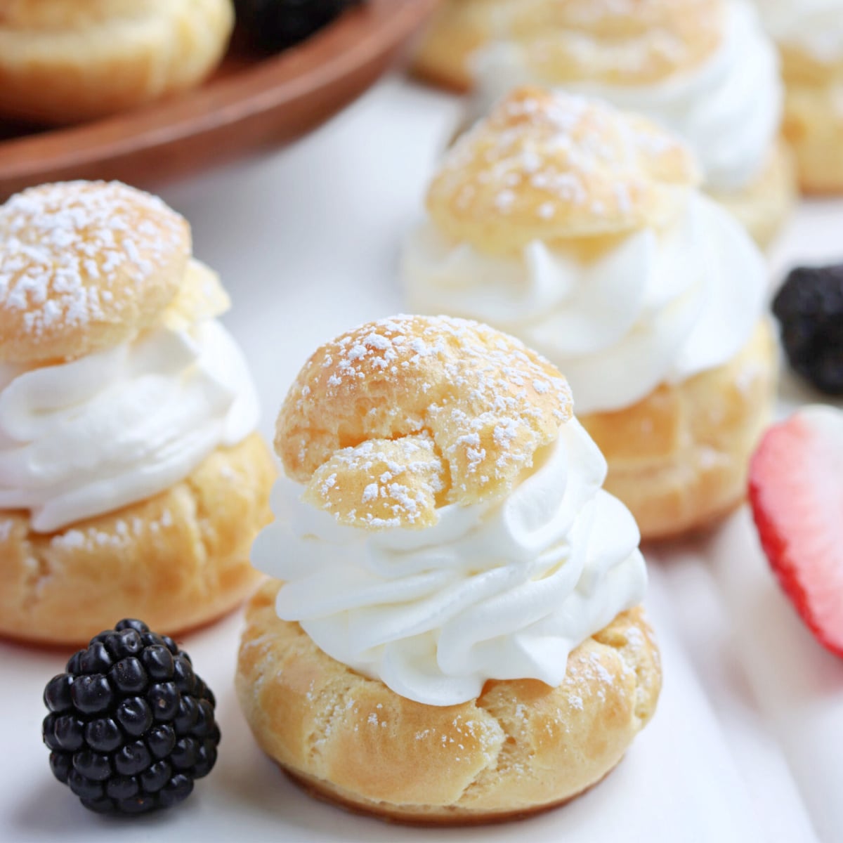 Cream puffs sprinkled with powdered sugar with fresh berries in the background. 