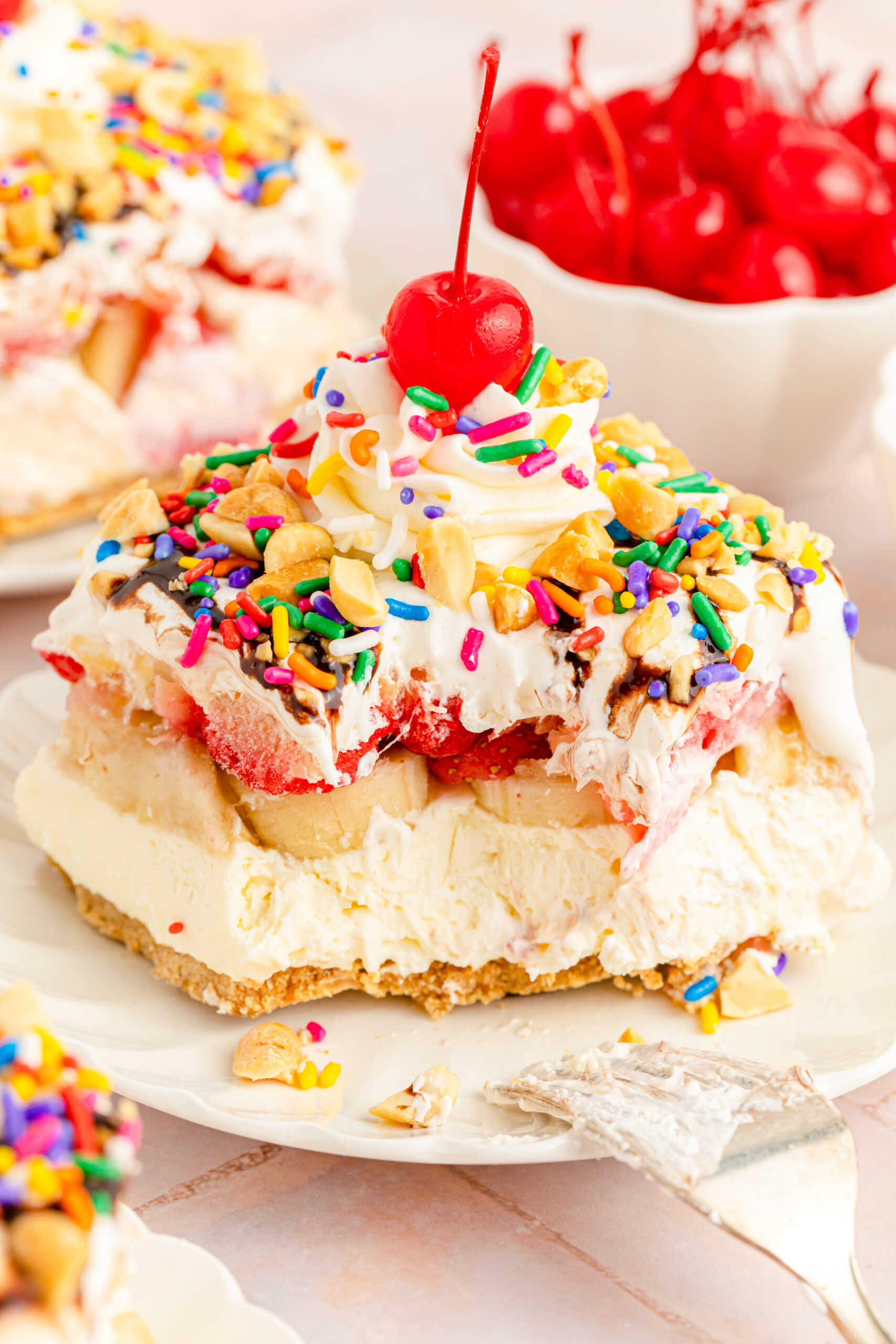 Close up view of a slice of No Bake Banana Split Cake on a white plate with a bite removed by a silver fork.