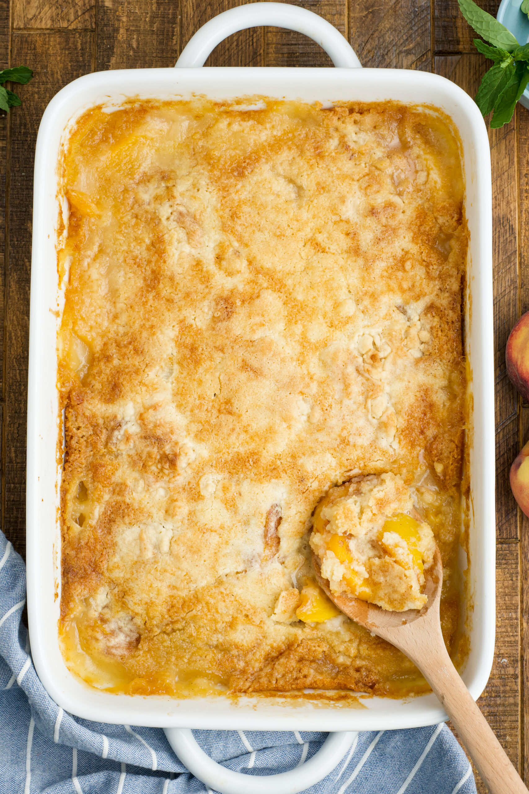 Overhead view of Peach Dump Cake in a white baking dish with a wooden spoon removing a serving.