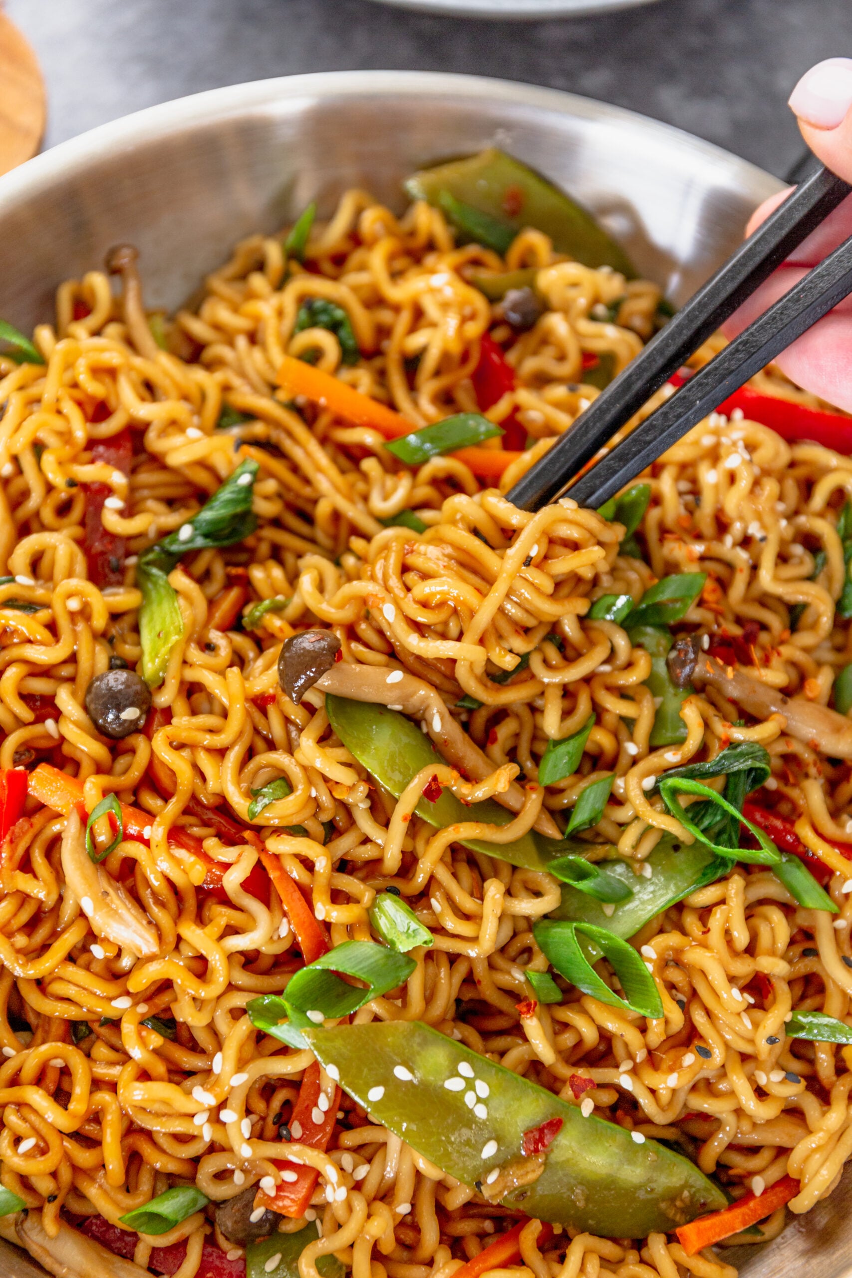 A pair of chop sticks removing Ramen Stir Fry from a silver frying pan.