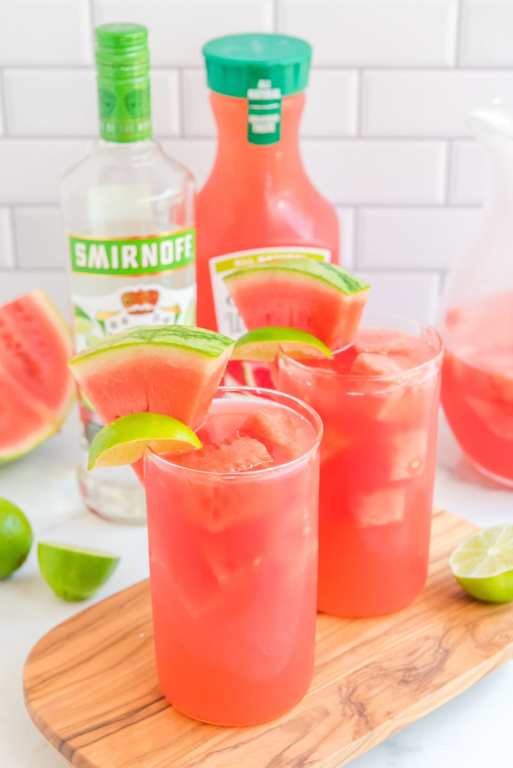 Front view of Watermelon Cocktail in two clear glasses garnished with lime and watermelon wedges on a wooden board.