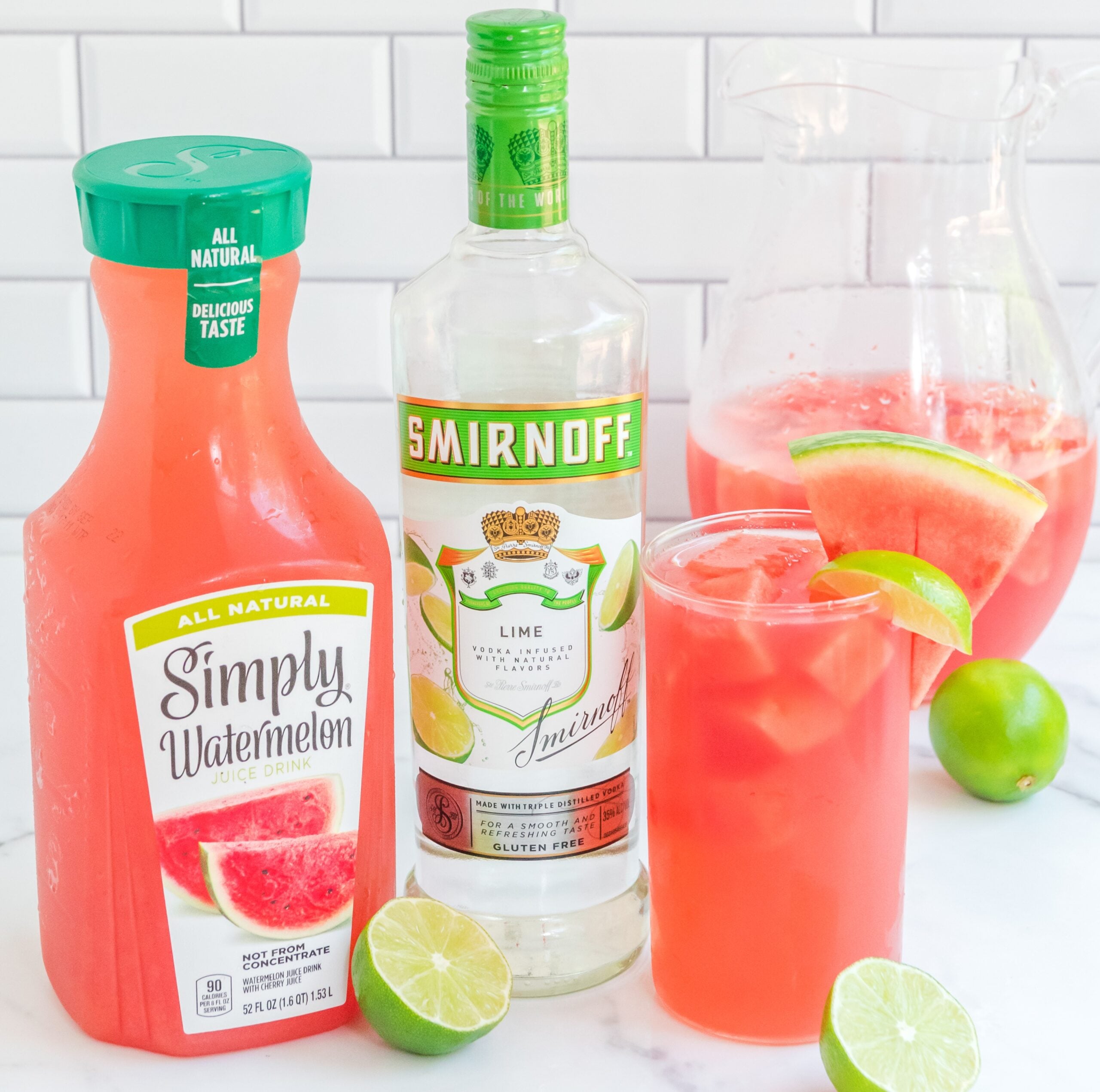 Front view of Watermelon Cocktail in a clear glasses garnished with lime and watermelon slices next wo watermelon juice bottle and a vodka bottle