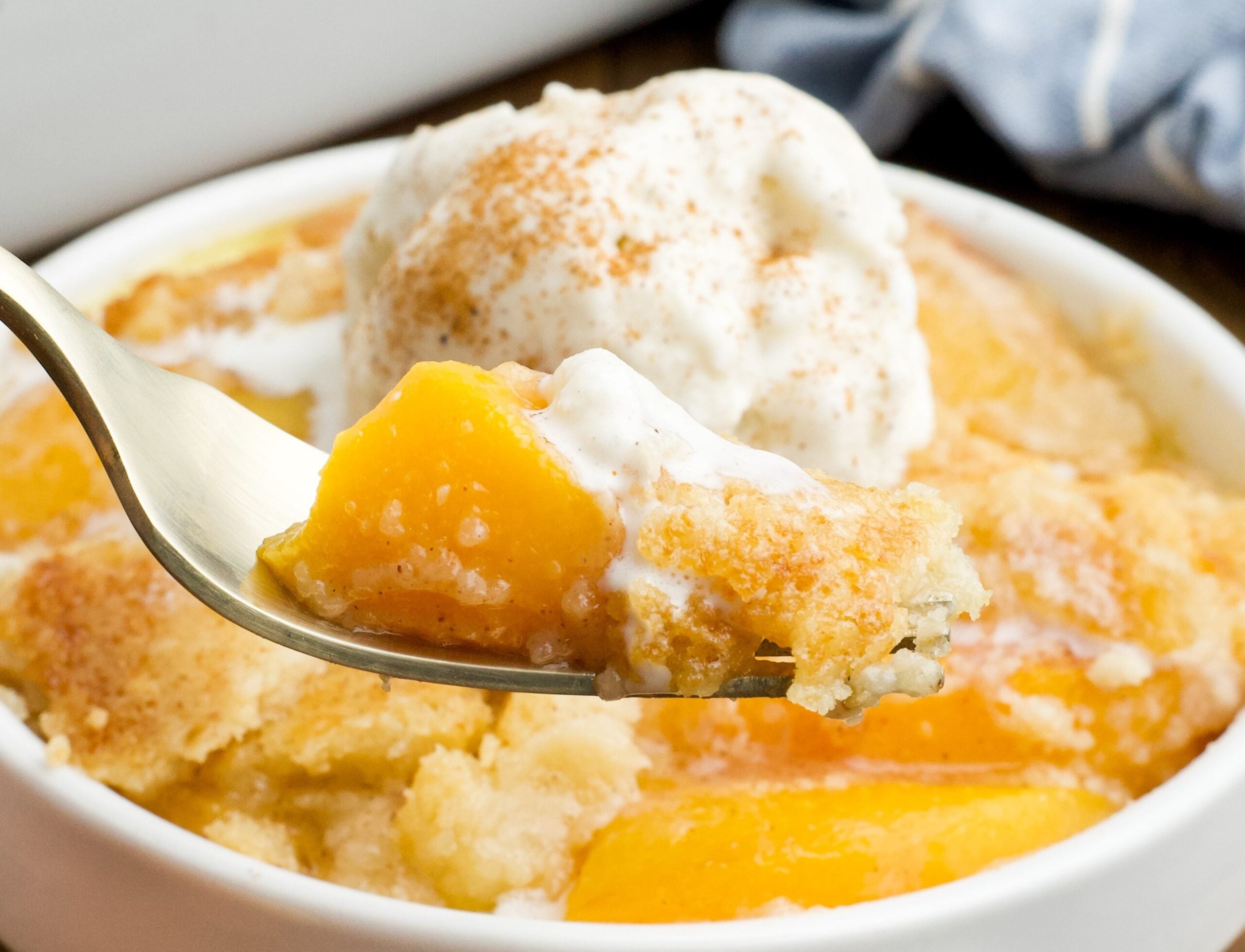 close up of of a serving of Peach Dump Cake in a white bowl with a gold fork removing a bite.