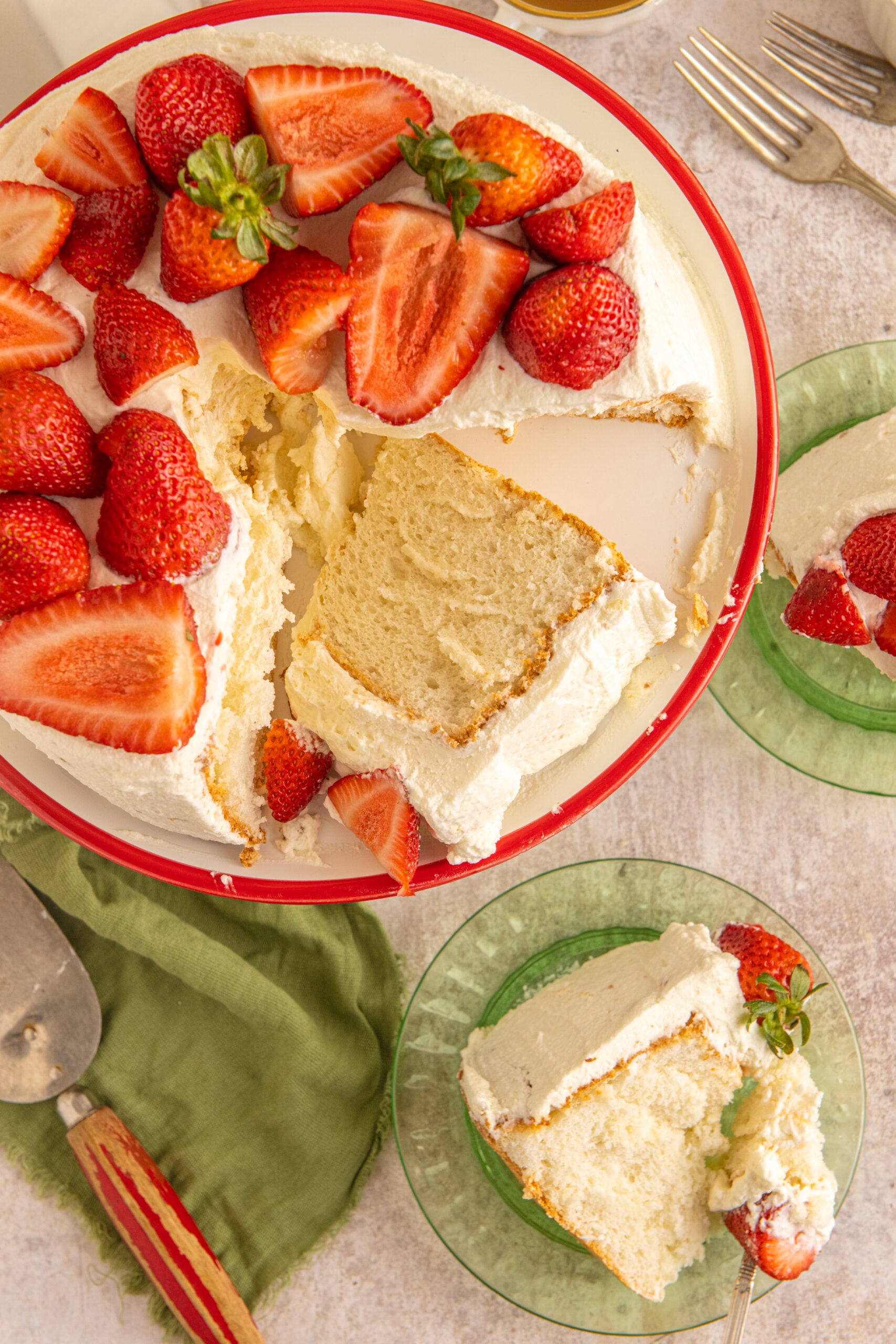 Overhead view of a cut piece of Strawberry Angel Food Cake on a white and red cake stand with two pieces served on green plates in the background.
