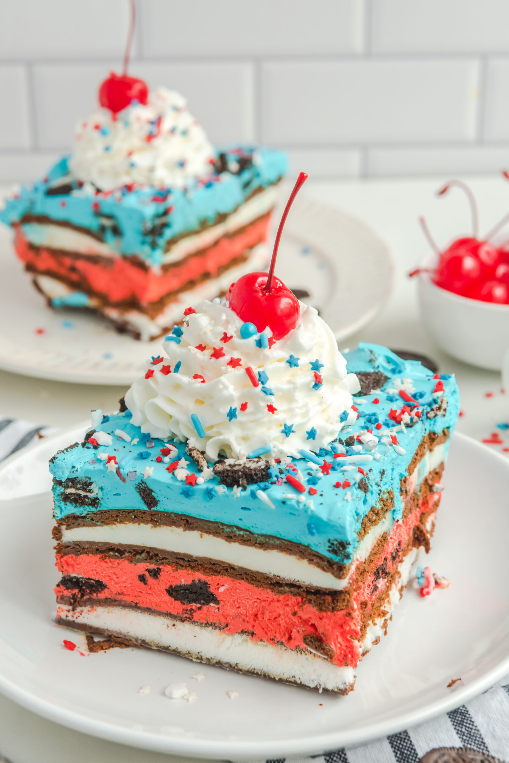 Front view of a slice of Patriotic Ice Cream Sandwich Cake topped with whipped cream and cherry.