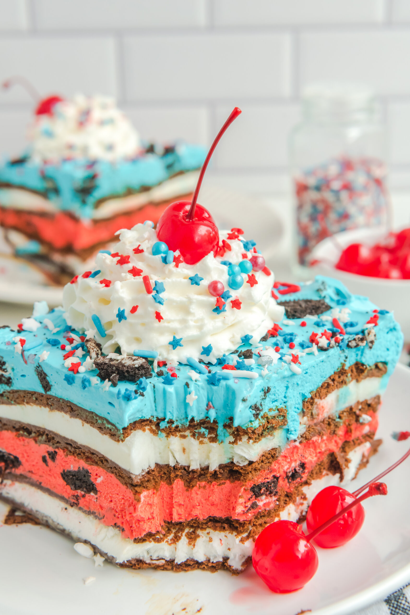Front view of a slice of Patriotic Ice Cream Sandwich Cake with a bite missing topped with whipped cream and cherry.
