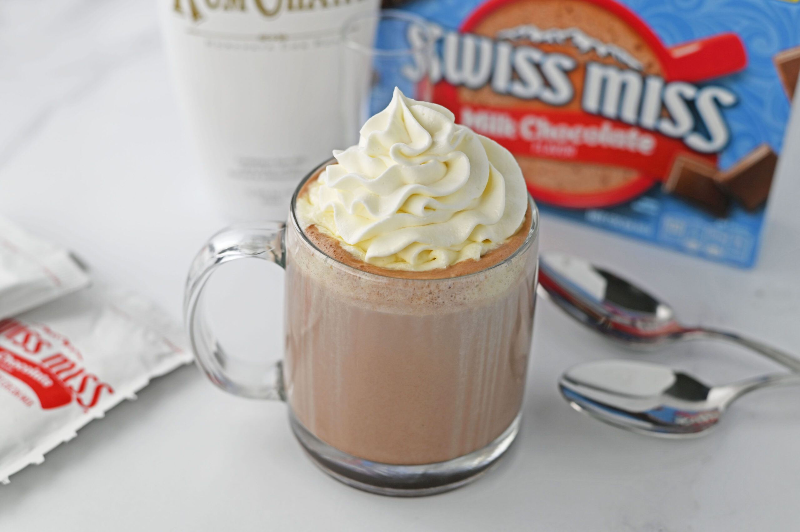 rumchata in a clear glass mug topped with whipped cream with a box of swiss miss and rumchata in the background