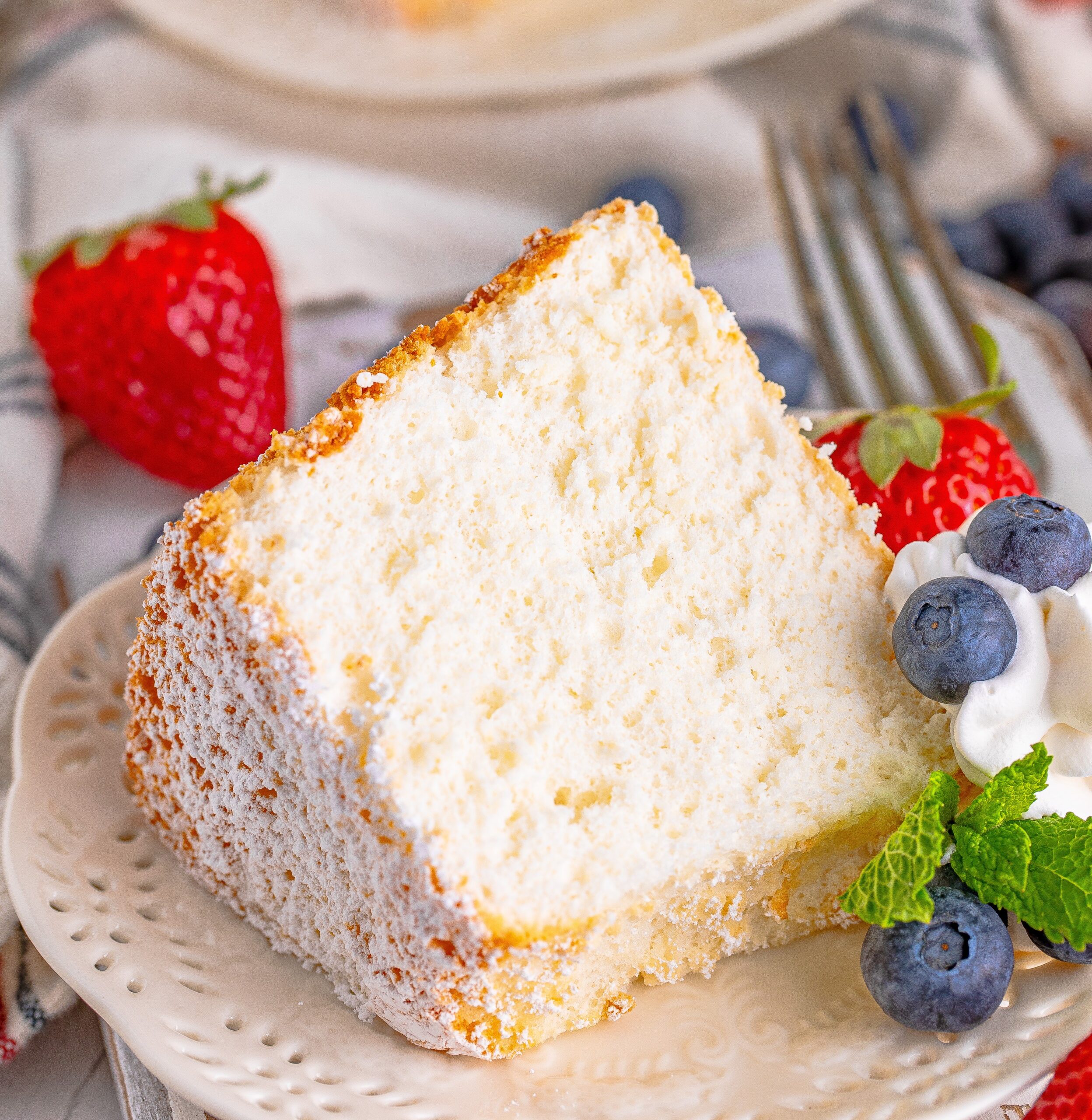 Close up of a slice of Angel Food Cake on a white plate with whipped cream, fresh berries, and mint