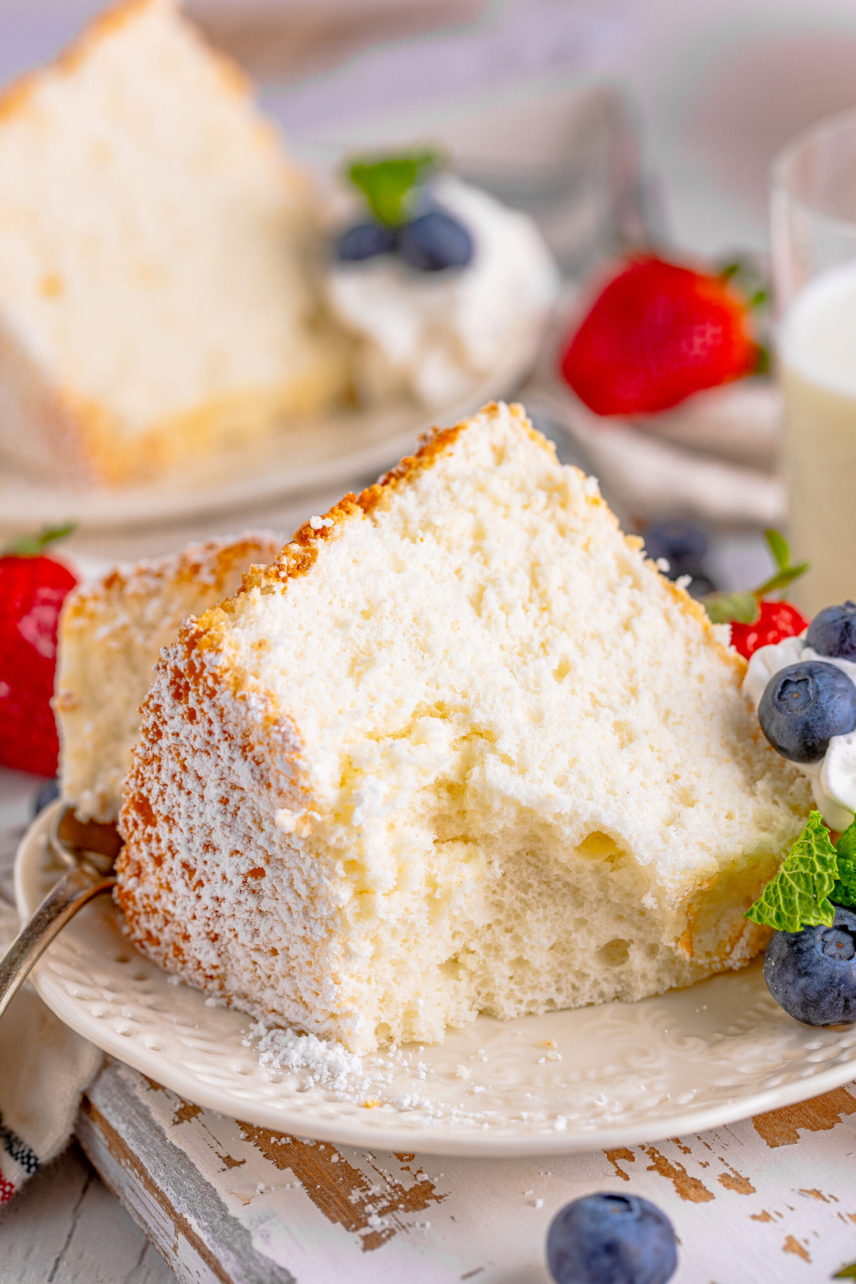 Close up of a slice of Angel Food Cake with a bite taken out of the cake, on a white plate with whipped cream, fresh berries, and mint