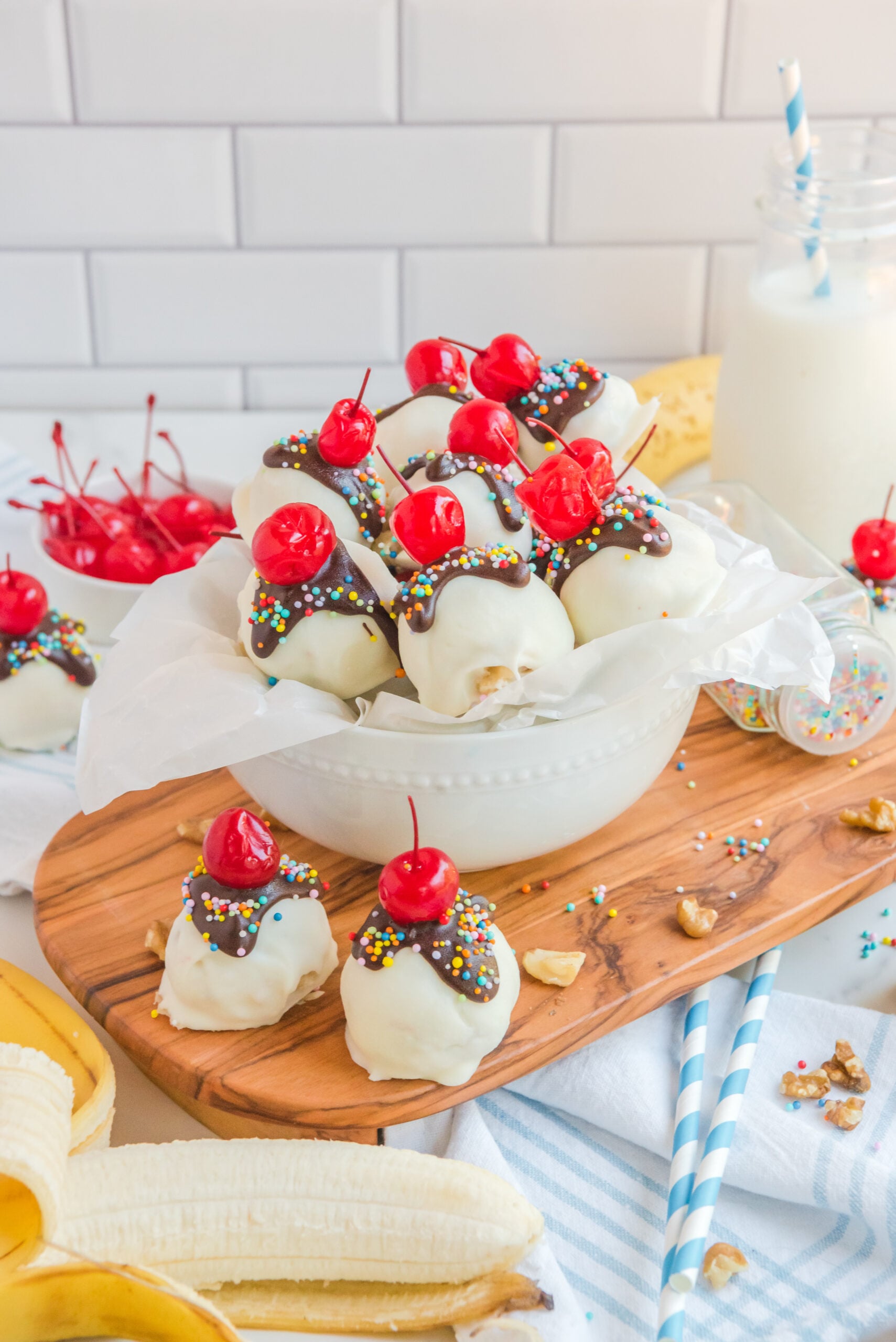 Front view of Banana Truffles on a wood cutting board surrounded by bananas, a bowl of cherries and nuts.