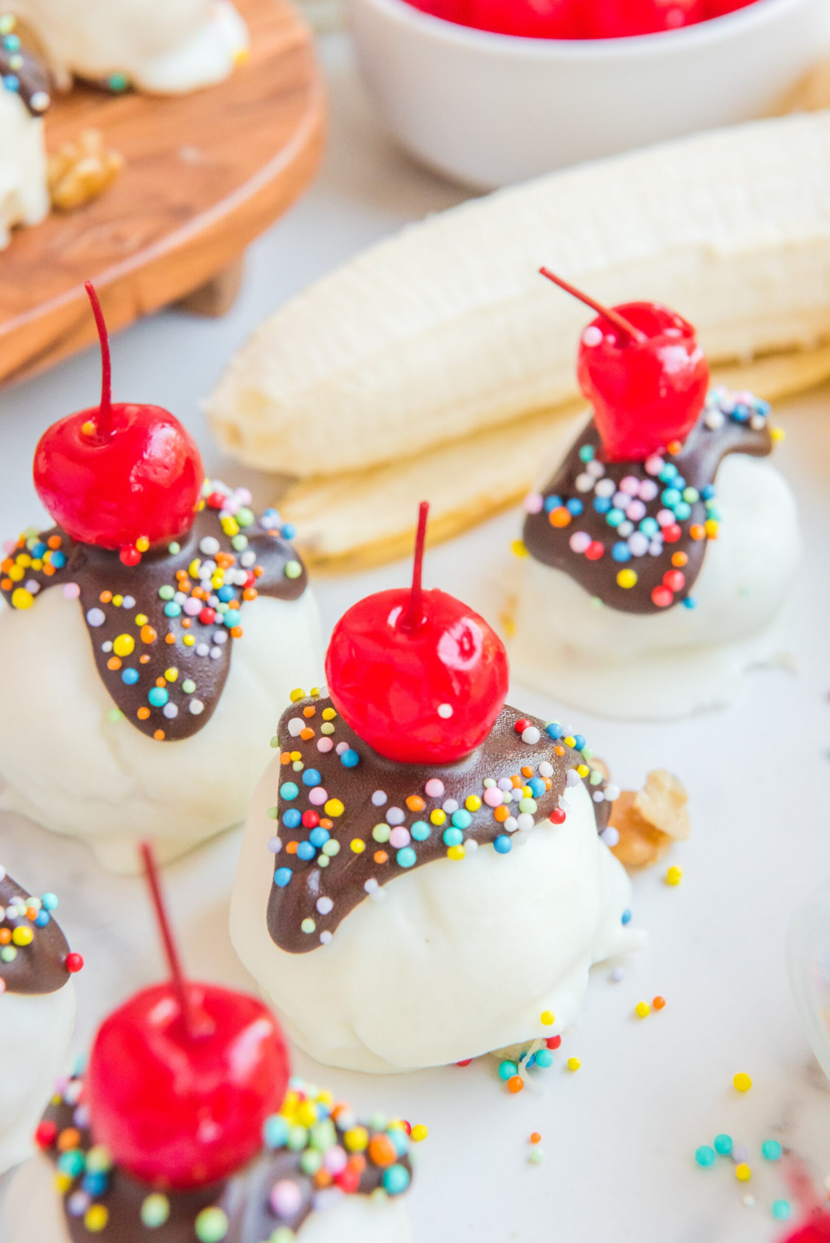 Front view of Banana Truffles surrounded by bananas, a bowl of cherries and sprinkles