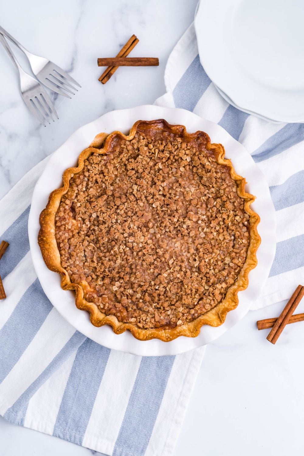 Apple Crumble Cheesecake Pie on table with cinnamon sticks and towel by pan 