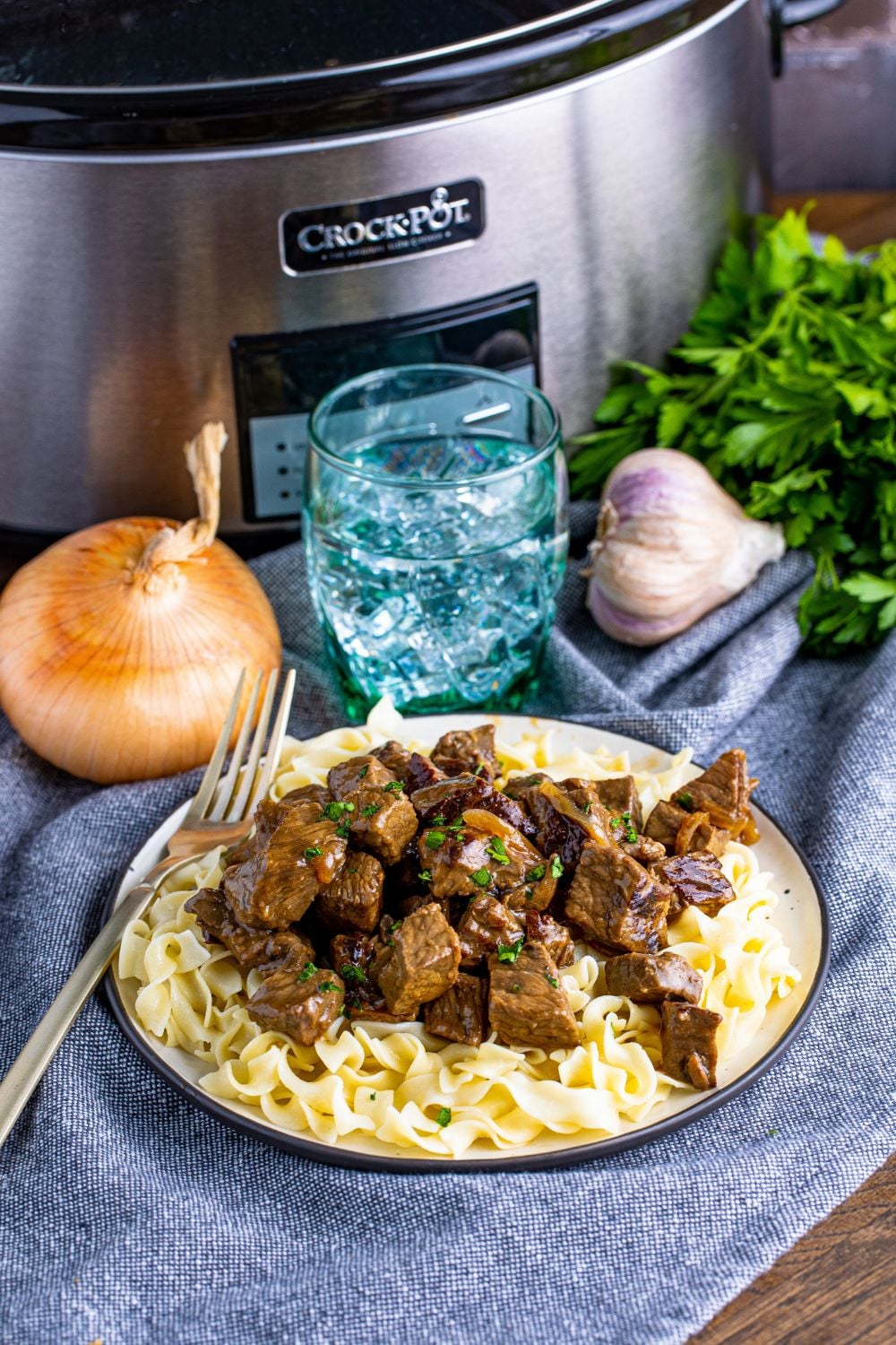 staged photo of crockpot in back, onions and garlic, and a plate with steak bites on it and a fork on side