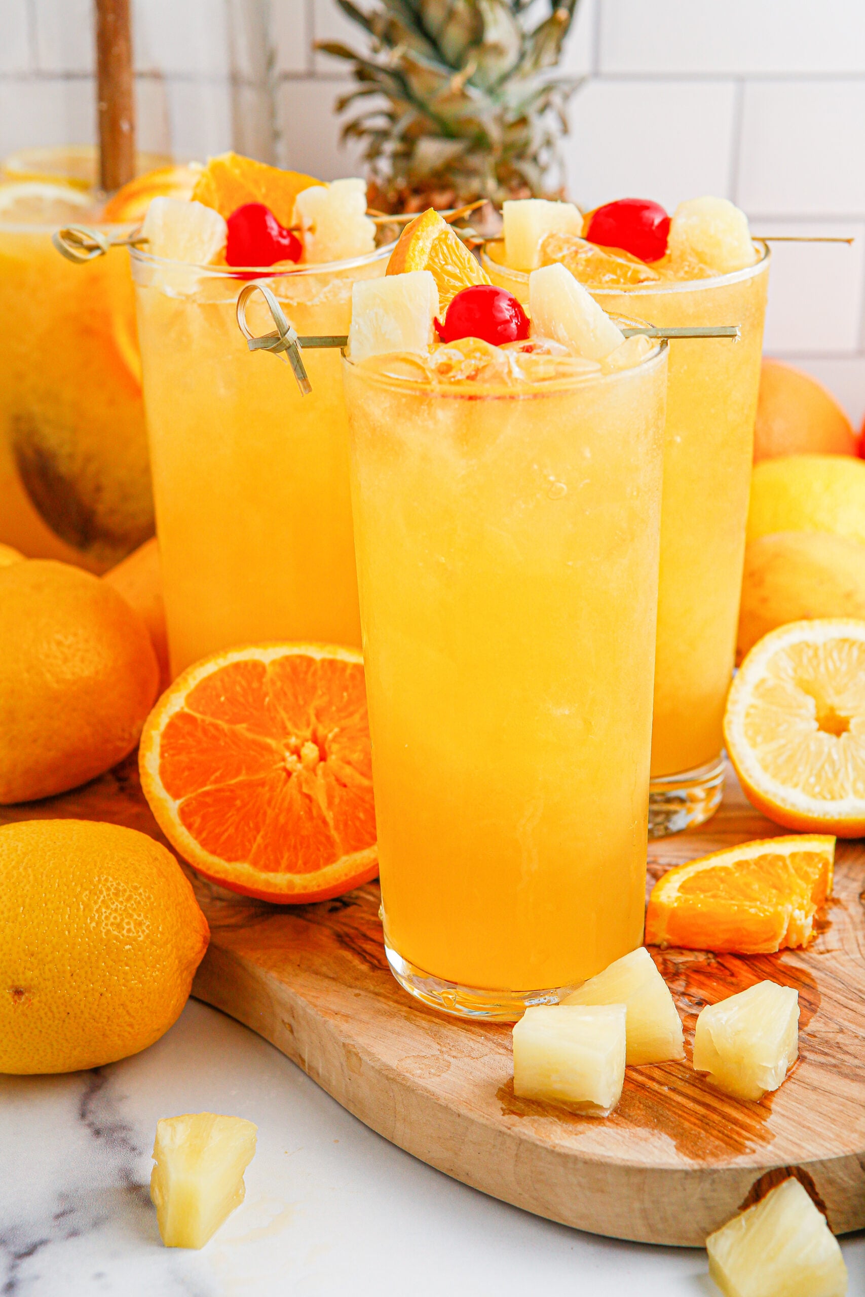 close up of the Tropical Lemonade Punch in 3 tall clear glasses on a wooden table with fruit around it and a pineapple in the background 