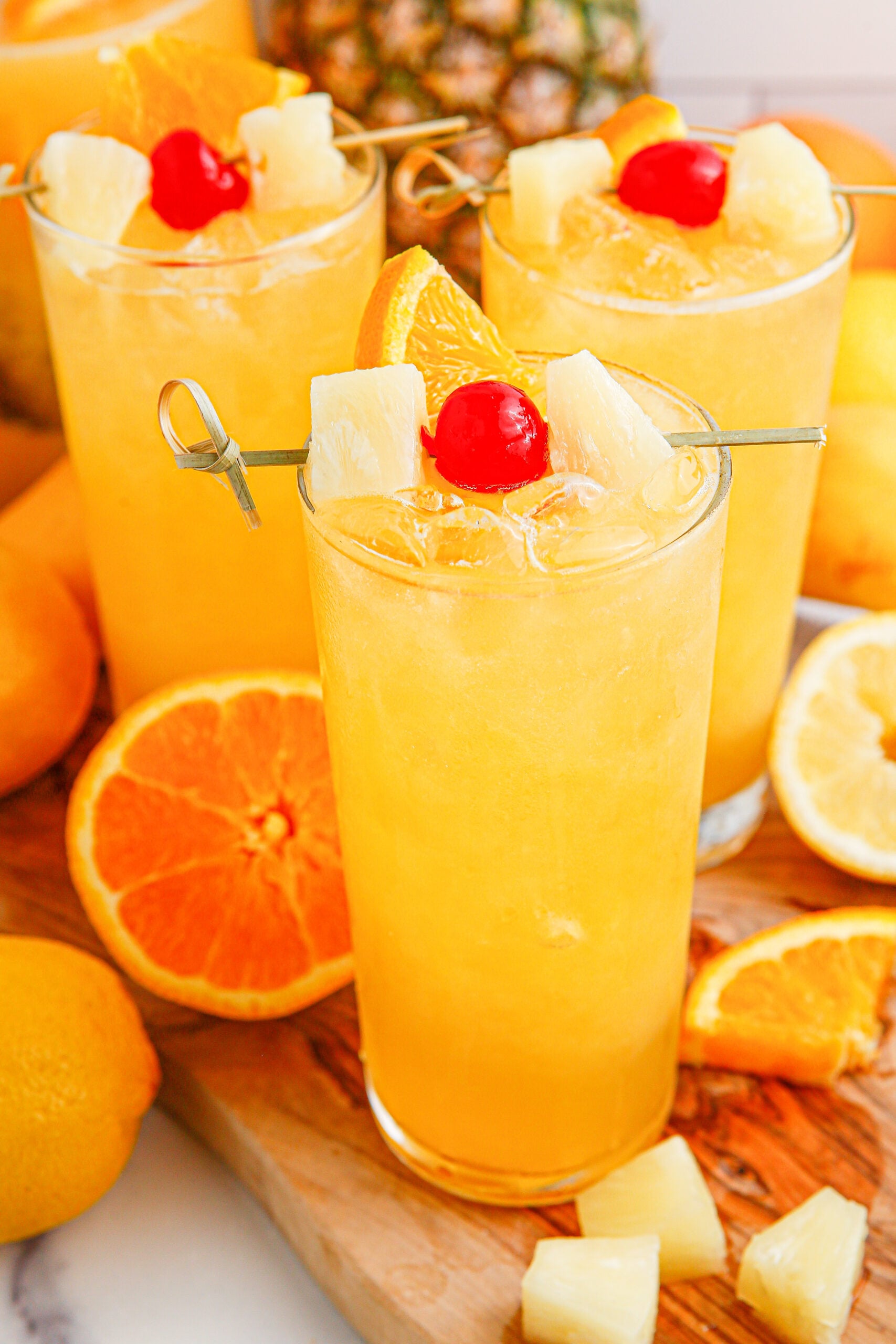 Tropical Lemonade Punch in 3 tall clear glasses on a wooden table with fruit around it and a pineapple in the background 
