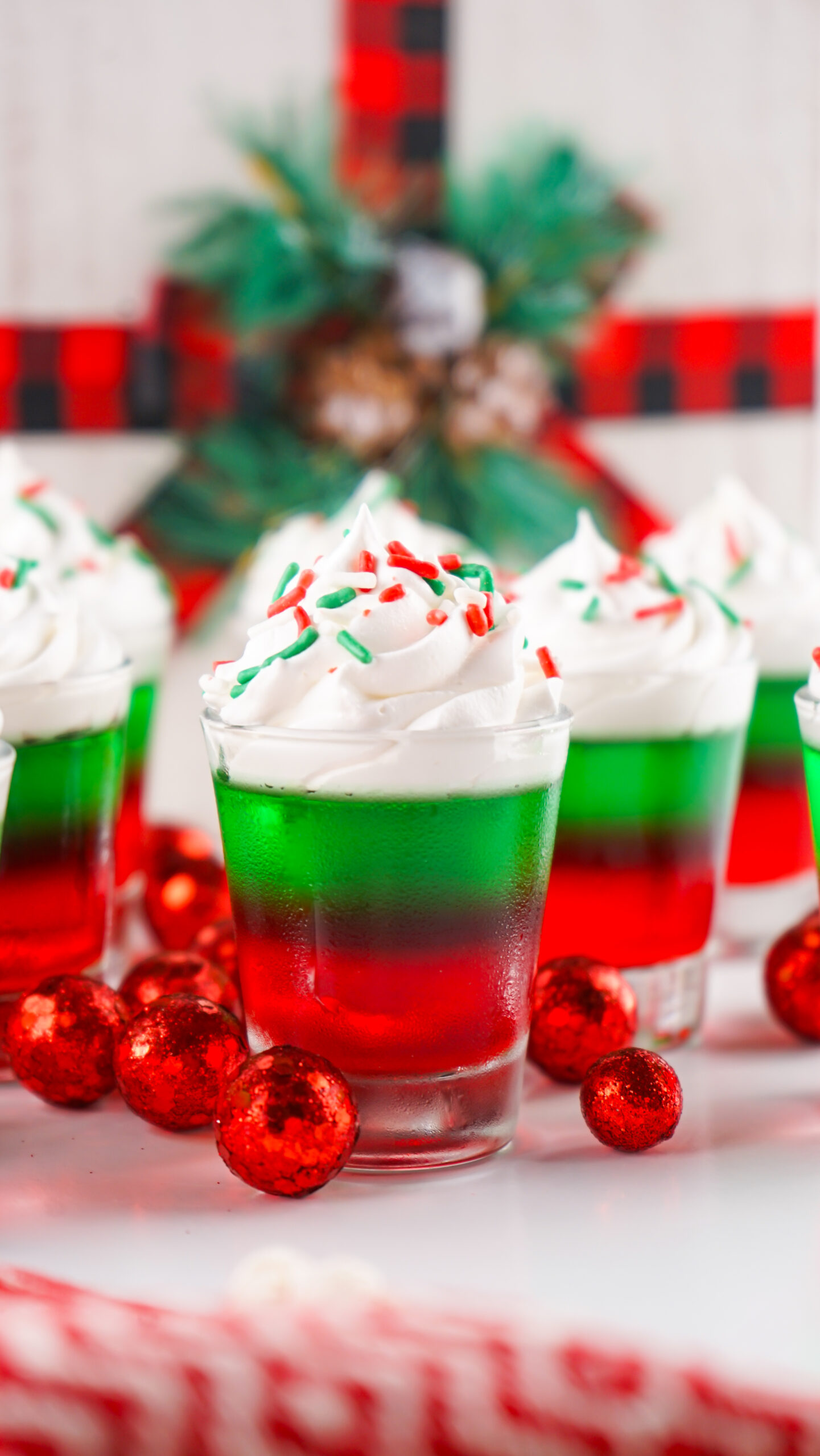 Christmas Jello Shots on a white able with red ornaments around them topped with holiday sprinkles and whipped cream