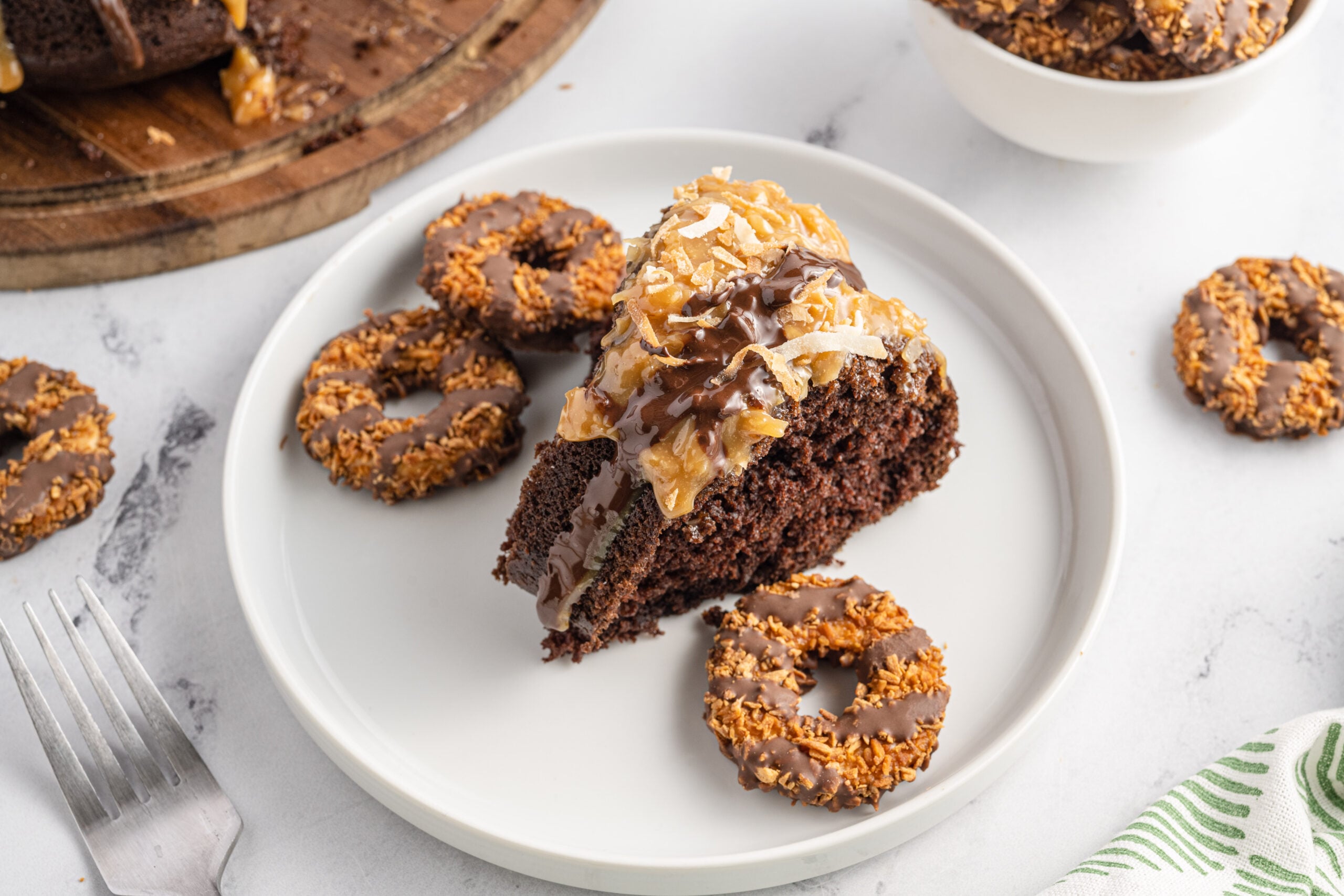 Close up view of a slice of Samoa Cake on a white plate with samoa cookies next to it