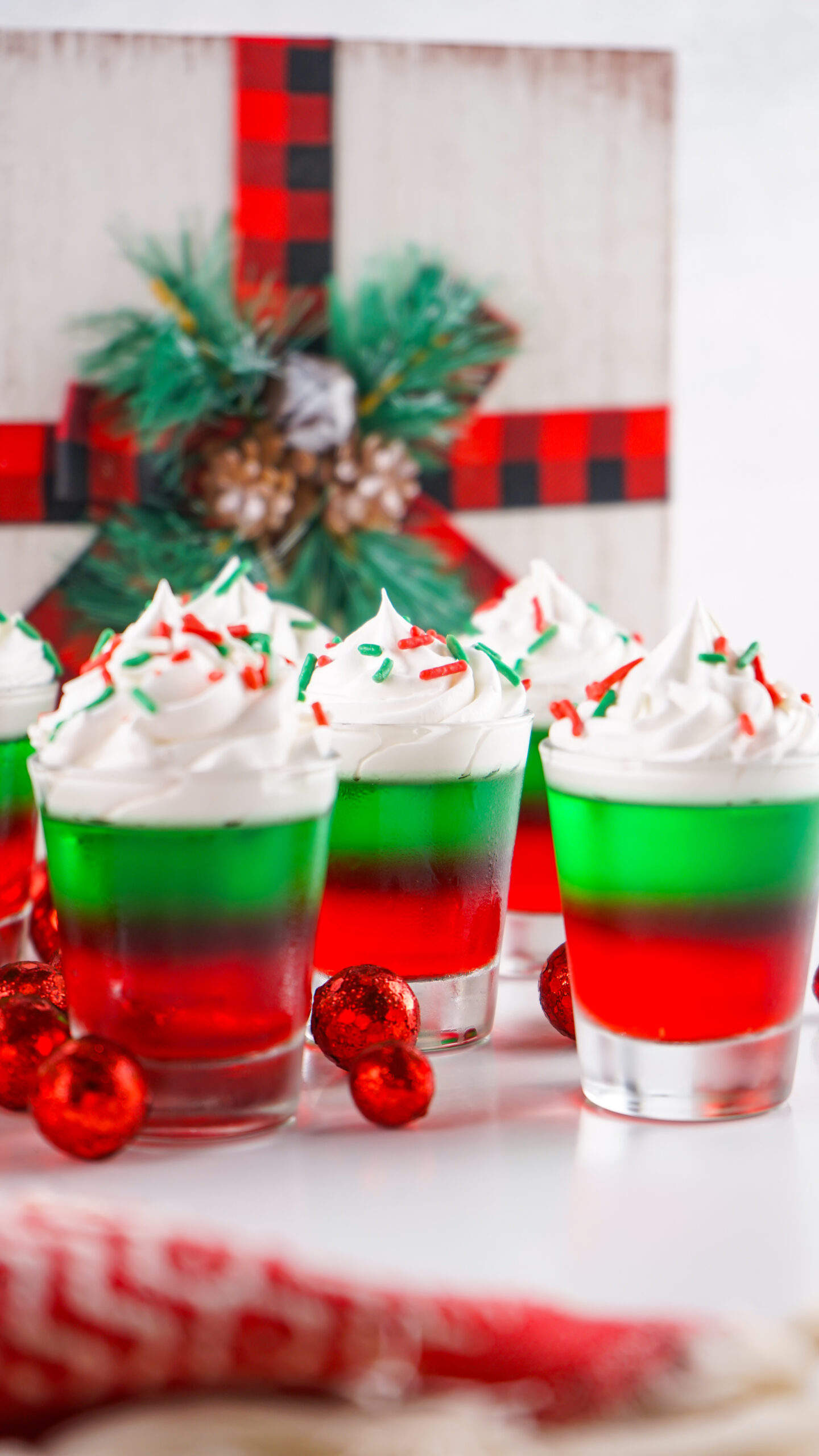Christmas Jello Shots on a white able with red ornaments around them topped with holiday sprinkles and whipped cream