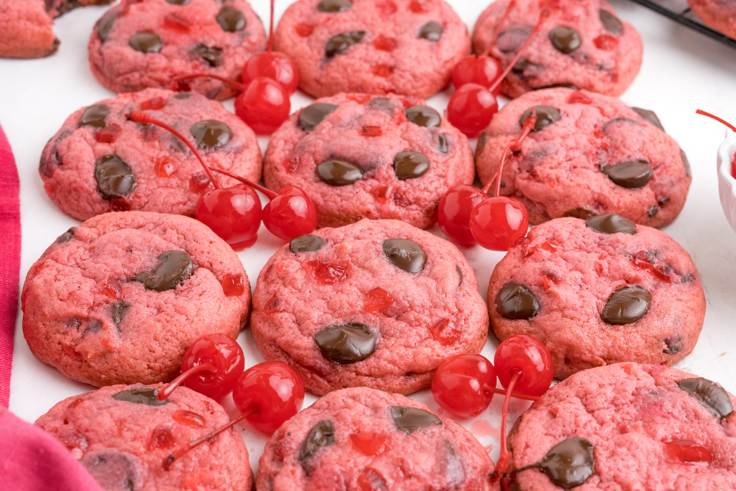 Cherry Chocolate Chip Cookies placed on table with maraschino cherries in between cookies on table to offer color and cookies in a single line 