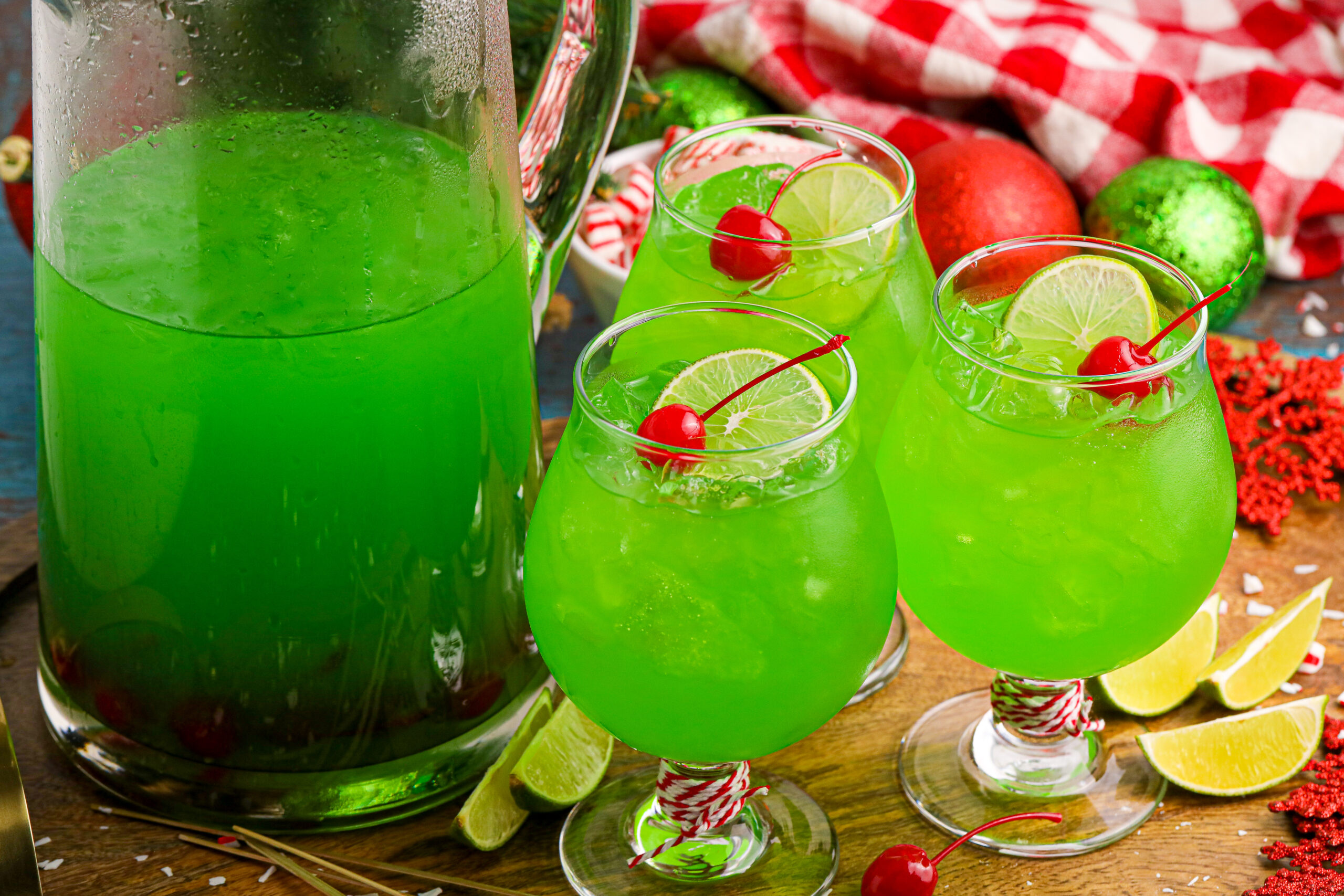 Grinch Punch in a clear glasses on a table with a cherry and a lime slice in the glasses, next to a large pitcher