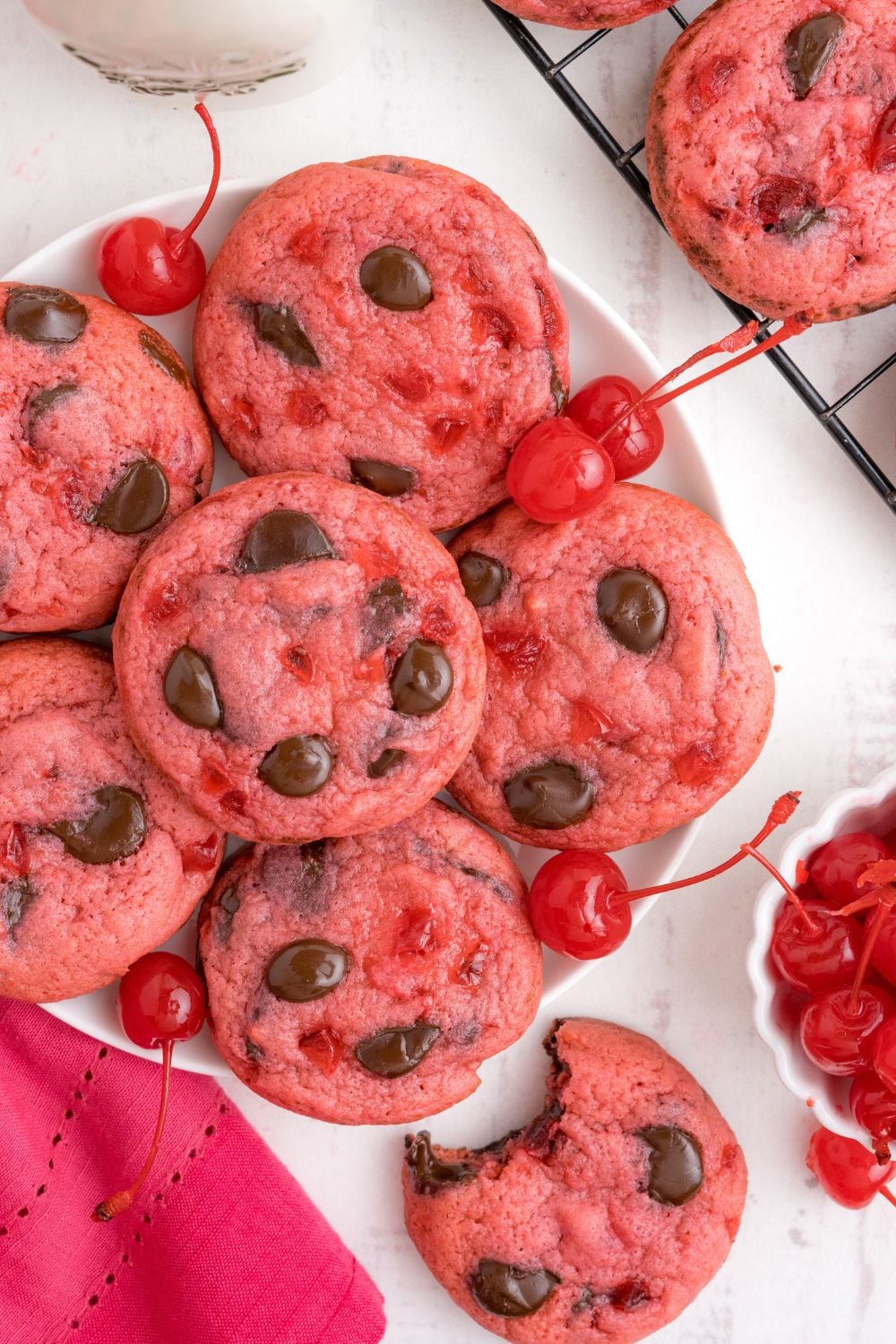 Cherry Chocolate Chip Cookies on a plate with one cookie with a bite taken out placed on table and a bowl of cherries on side 