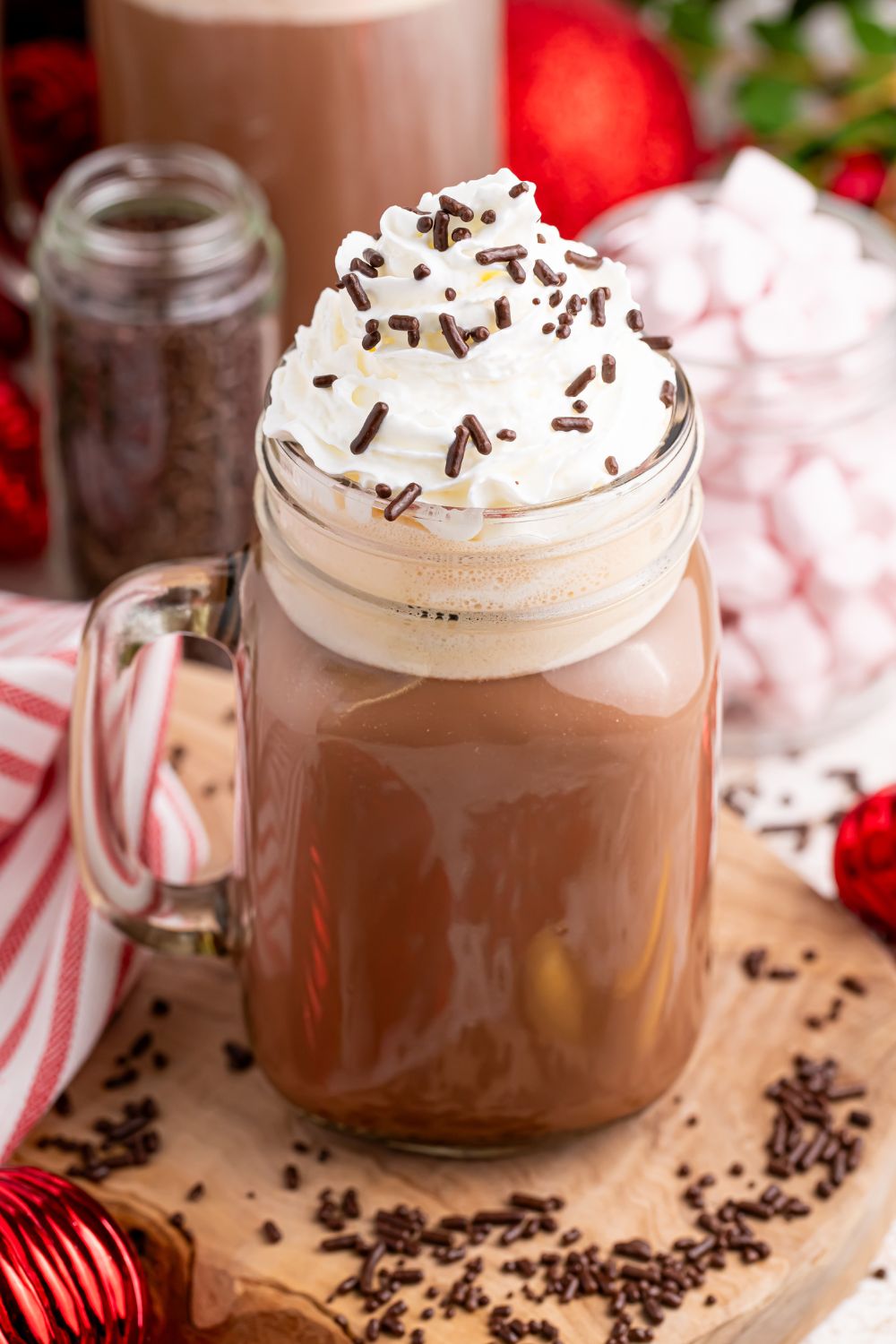 Dirty snowman cocktail in a glass on a wooden coaster with sprinkles and whipped cream on top. There is a red and white towel and other Christmas decor behind the glass
