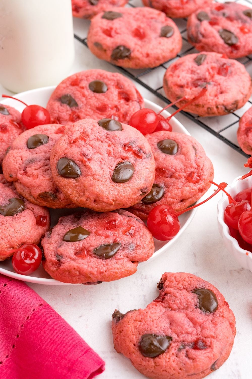 Cherry Chocolate Chip Cookies side view with a plate stacked of cookies and a tall glass of milk beind the plate 