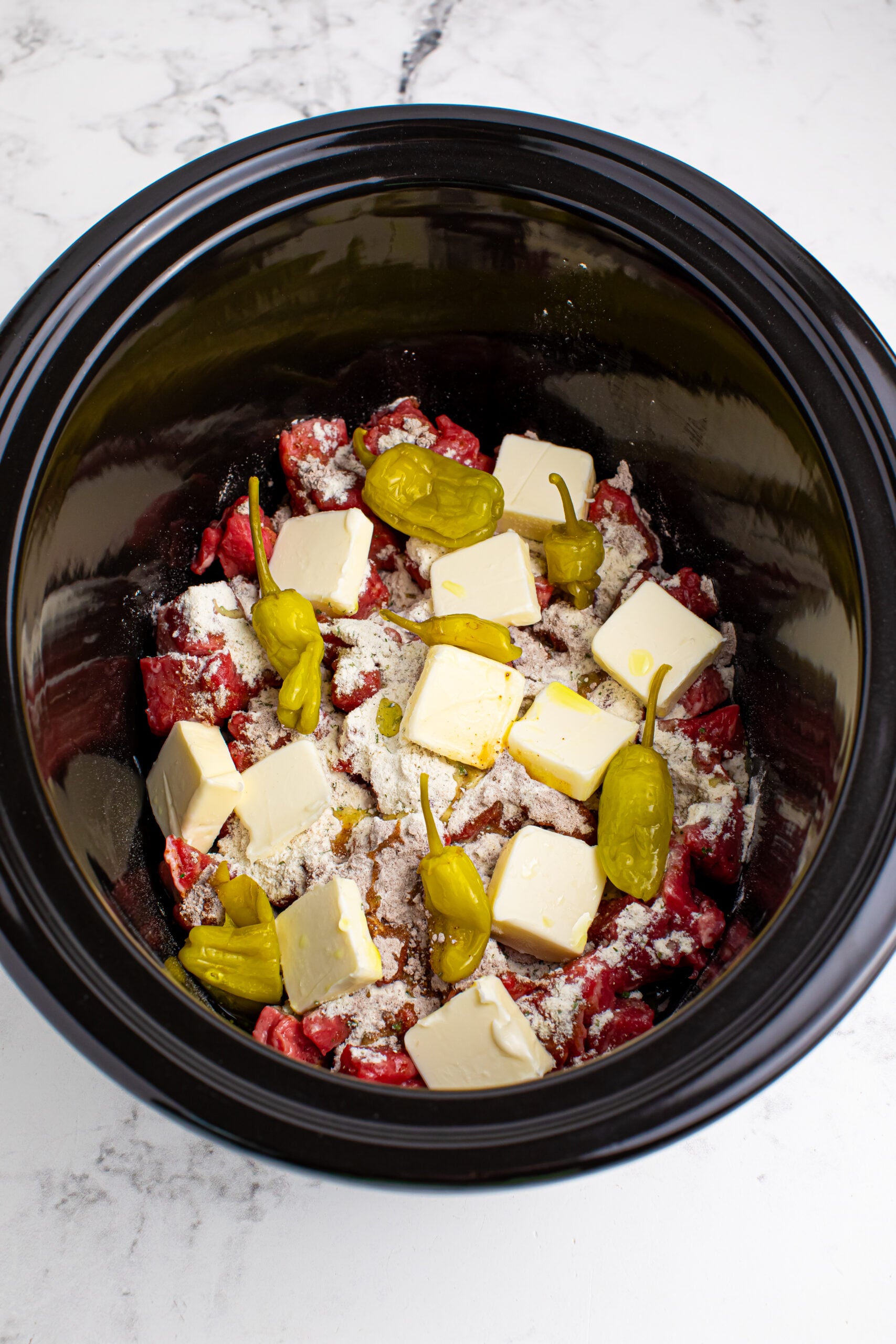 Crockpot Mississippi Steak Bites with diced steak and dry seasoning sprinkled on top of meat and cubes of butter on top and peppers
