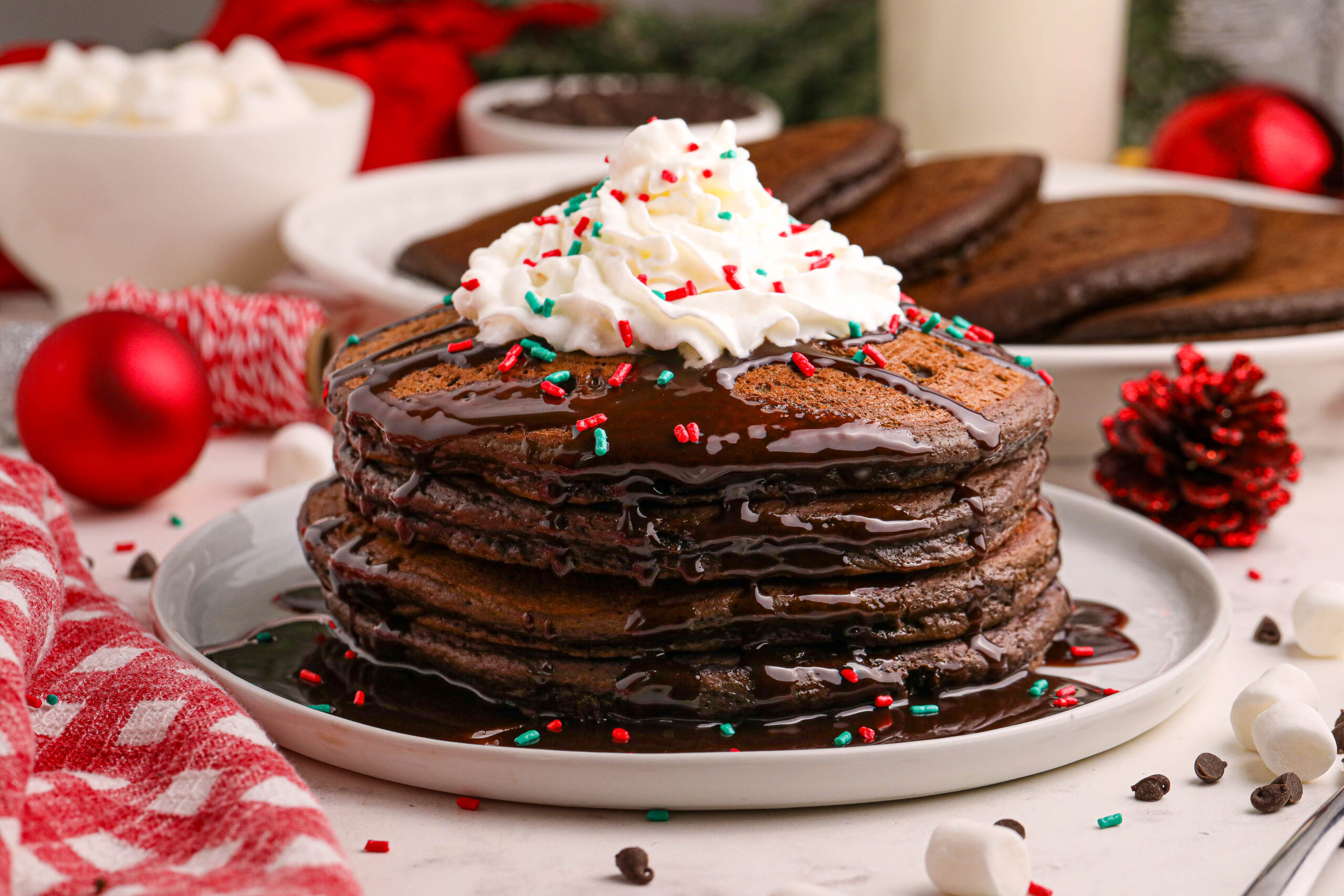 Hot Chocolate Pancakes stacked on a white plate topped with syrup and whipped cream and holiday sprinkles