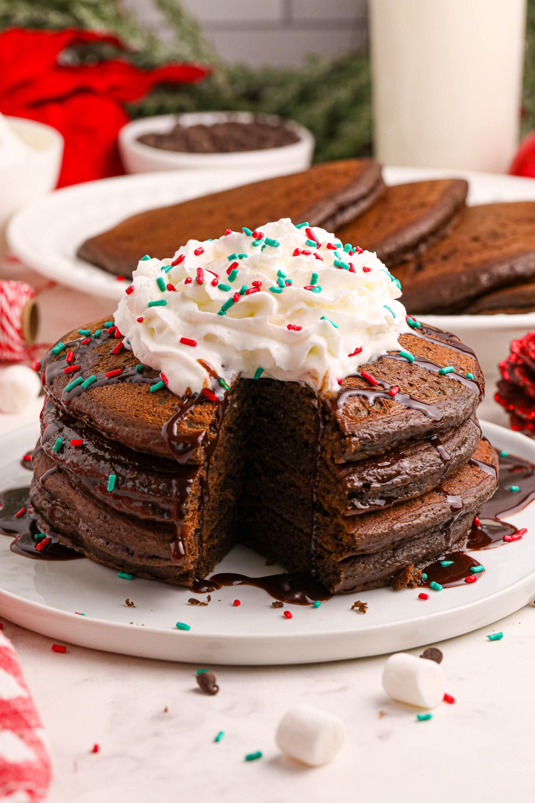 Hot Chocolate Pancakes stacked on a plate topped with syrup and whipped cream and holiday sprinkles with a bite removed