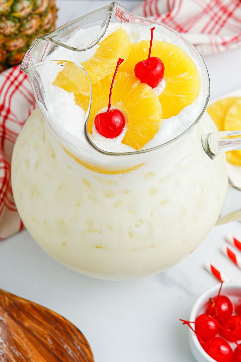 Pina Colada Punch in a pitcher with cherries and pineapple slices on the table and a wooden spoon 