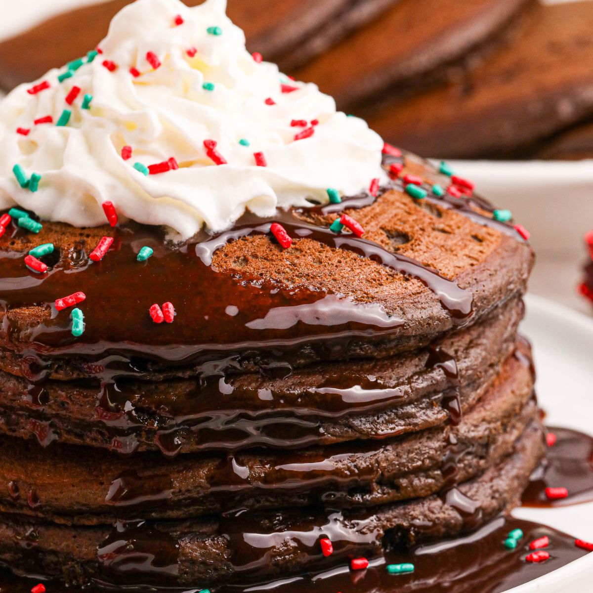 Hot Chocolate Pancakes stacked on a plate topped with syrup and whipped cream and holiday sprinkles