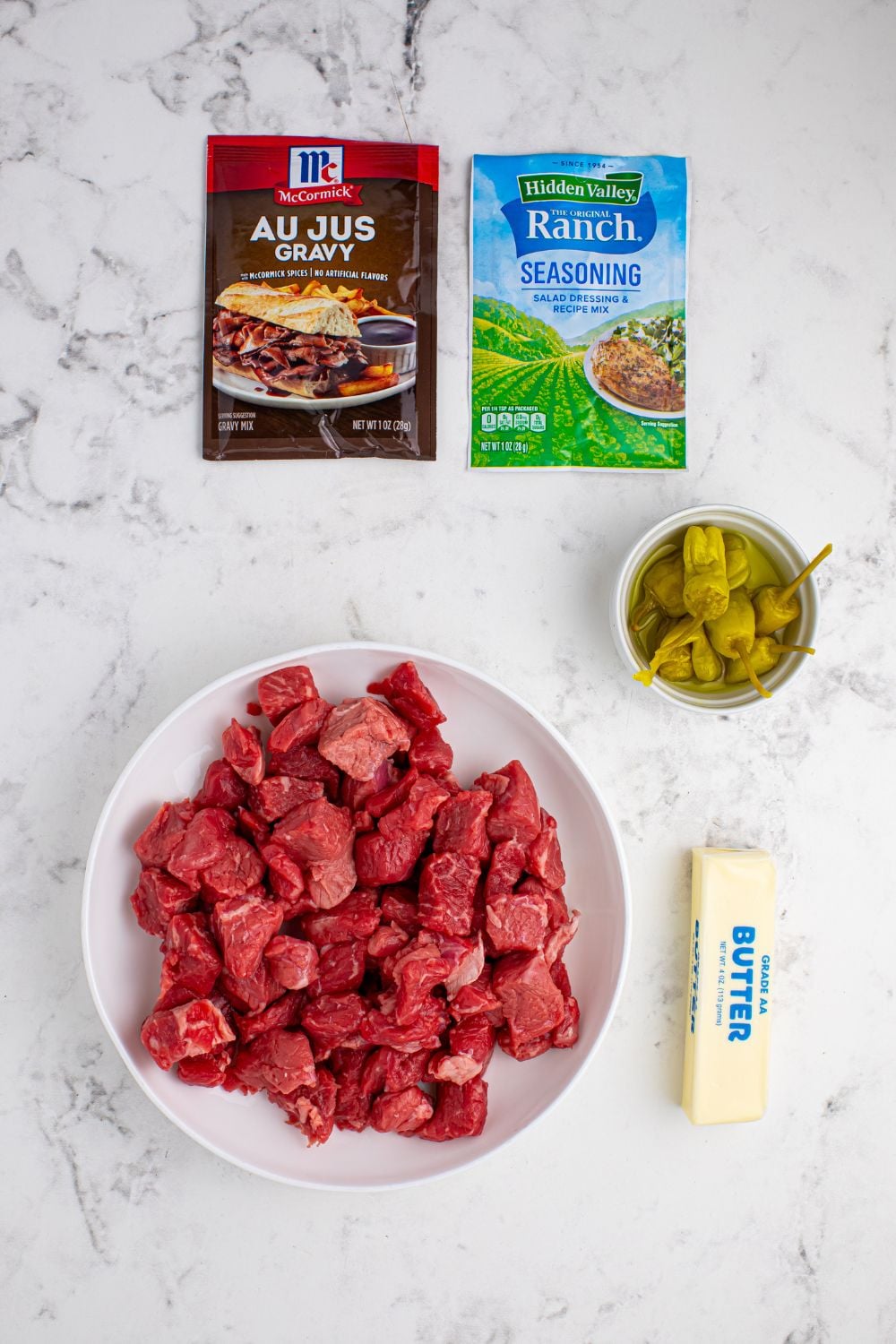 Crockpot Mississippi Steak Bites ingredients on table. Packet of au jus gravy, ranch seasoning, steak bites in a bowl, peppers in a bowl, and stick of butter