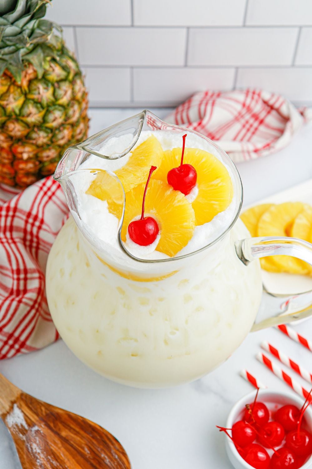 Pina Colada Punch in a pitcher on table with garnishes around the table, whole pineapple in background and cherries in a bowl 
