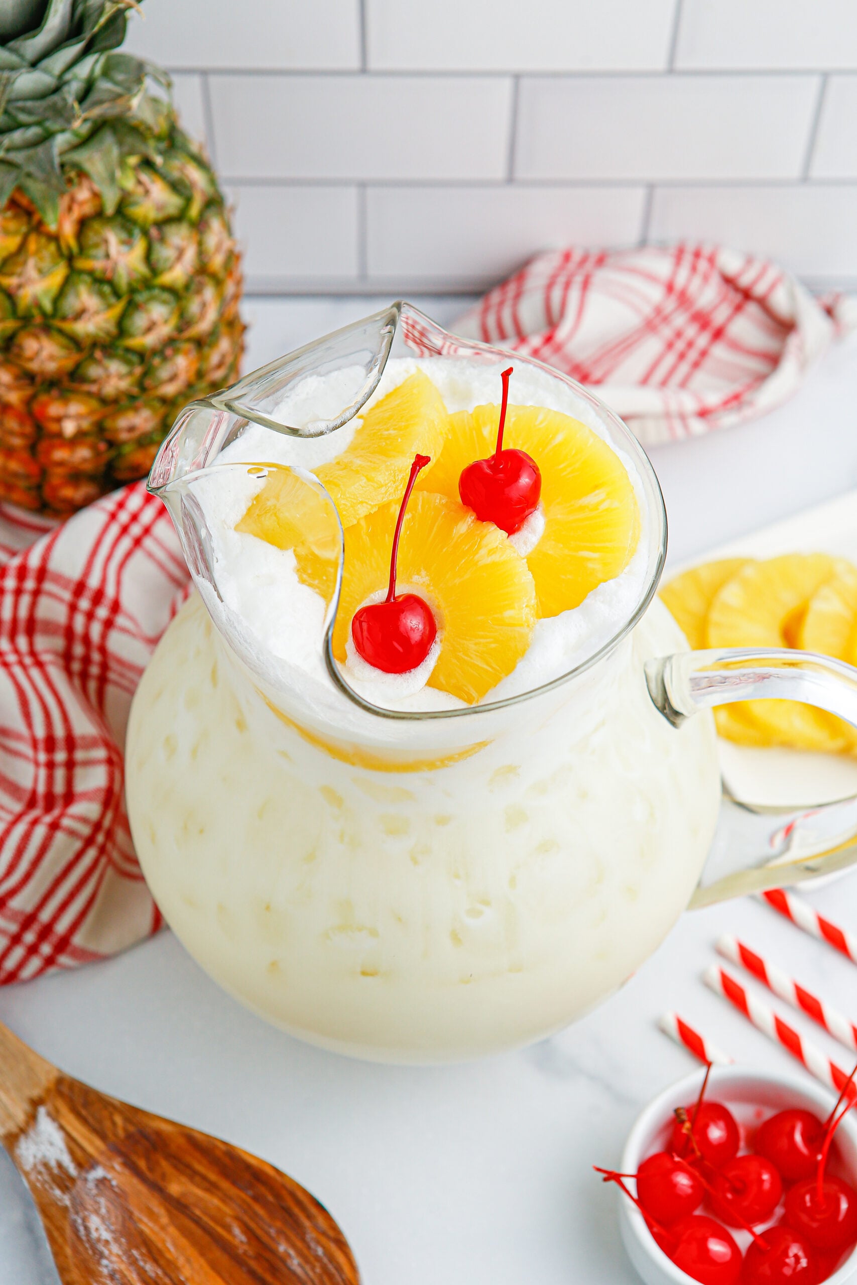Pina Colada Punch in a pitcher with cherries and pineapple slices on the table and a wooden spoon 