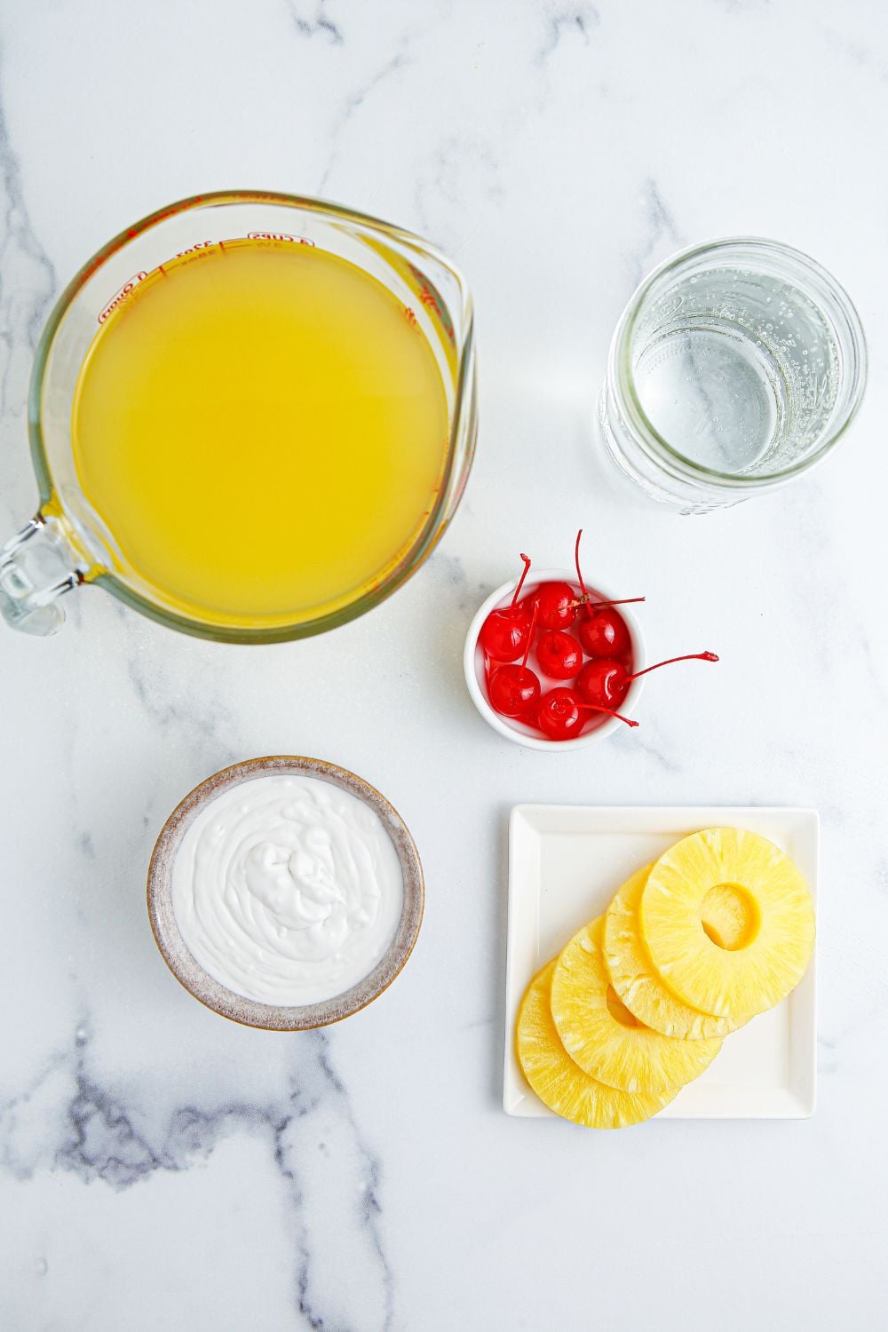 Pina Colada Punch ingredients on table. A pitcher of pineapple juice, coconut cream, sparking water, cherries, and pineapple wedges 