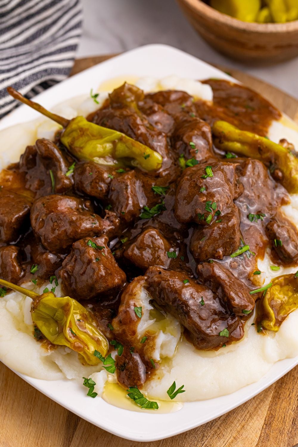 Crockpot Mississippi Steak Bites on a plate with mashed potatoes under the meat mixture and peppers in a bowl to the side